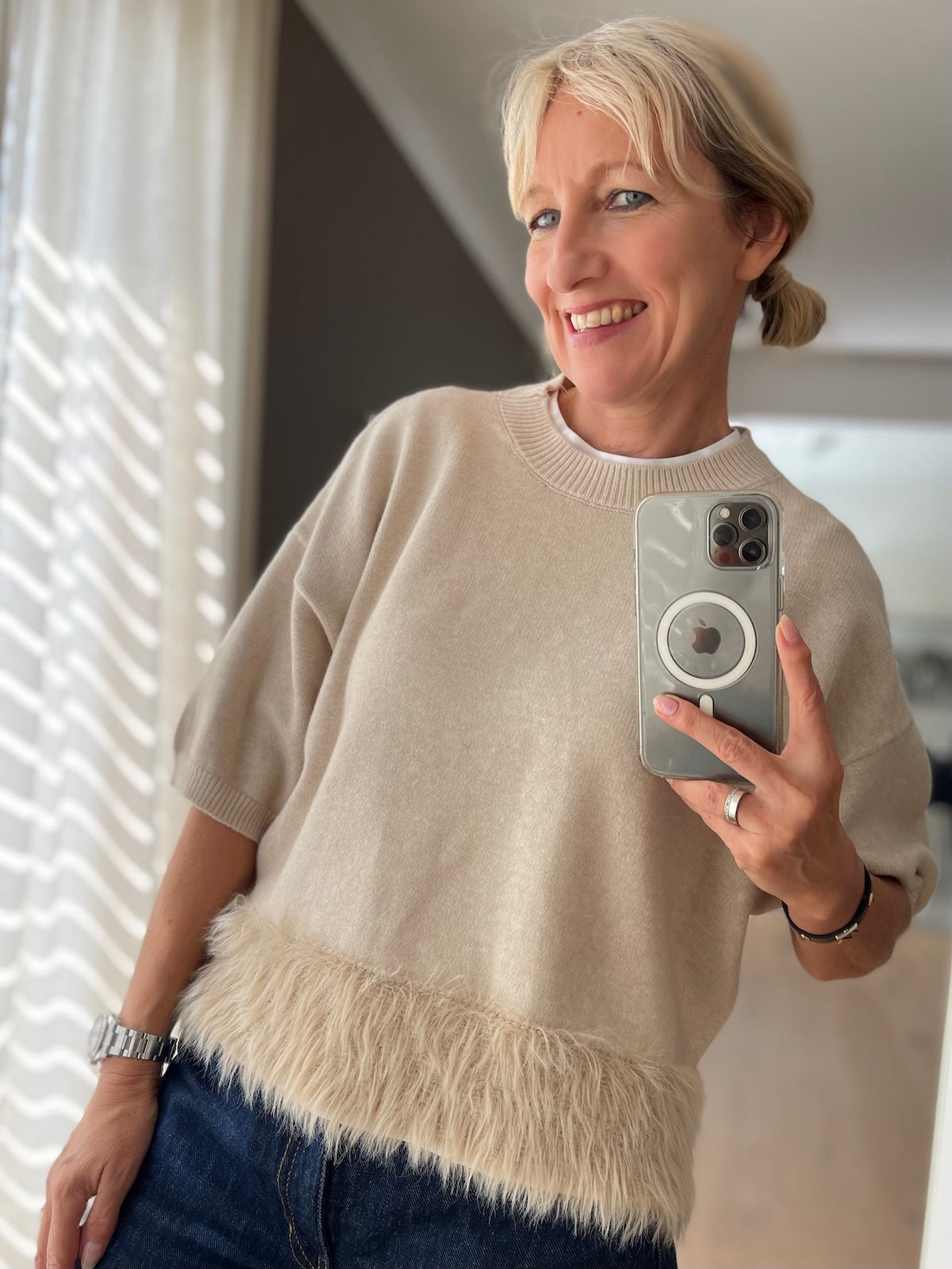 Alt-Text Bild 1: Blonde Frau trägt den beigen Boxy Strickpullover Franzy mit Fransen am Saum, modisch kombiniert mit einer dunkelblauen Jeans, fotografiert in einem hellen Raum.