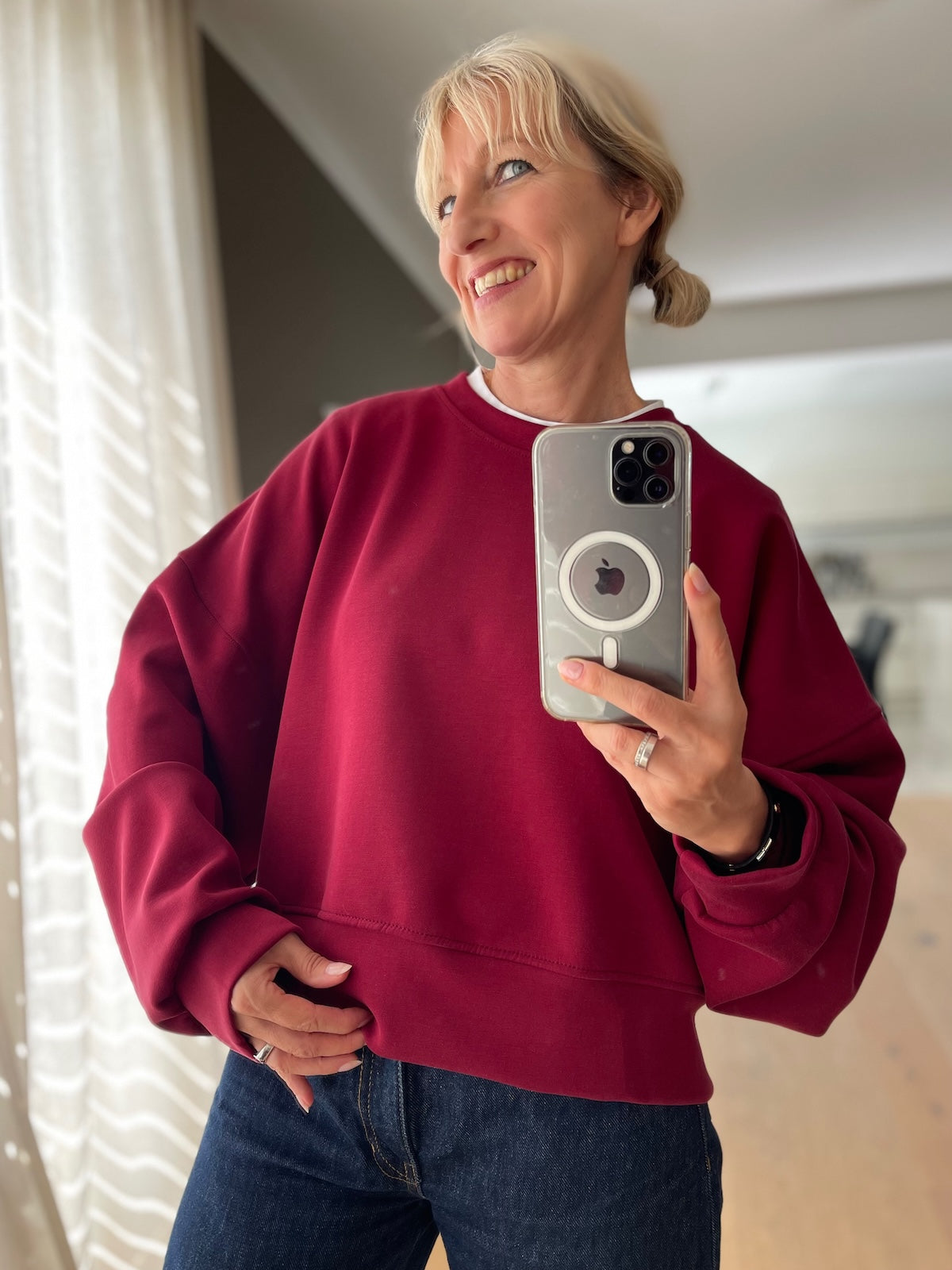 Blonde Frau präsentiert lächelnd das beerenfarbene Boxy Sweatshirt Romy mit überschnittenen Schultern und trendigen Fledermausärmeln, modisch kombiniert mit einer dunkelblauen High-Waist Jeans.