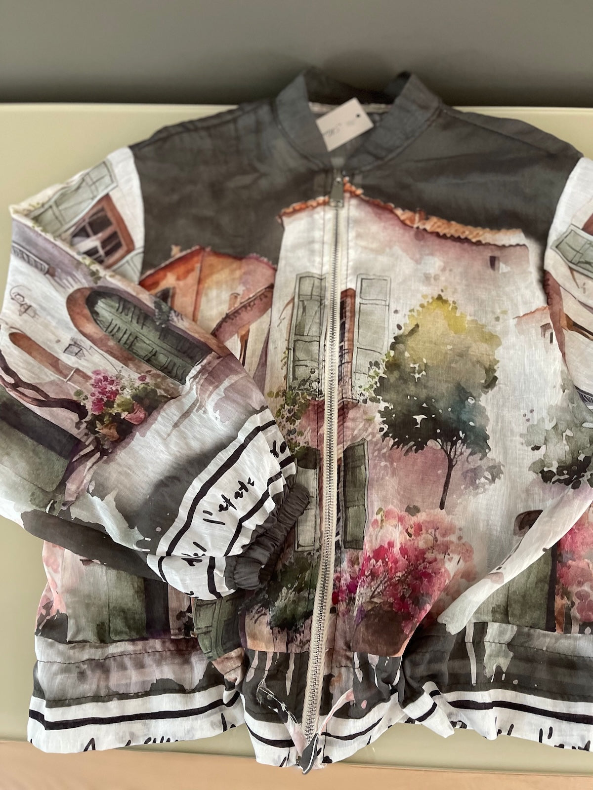 Flach liegende Detailaufnahme der ungefütterten Blouson-Jacke in Khaki. Der malerische Art-Print, der silberfarbene Reißverschluss und das Zugband am Saum sind auf dem transparenten Stoff gut erkennbar.