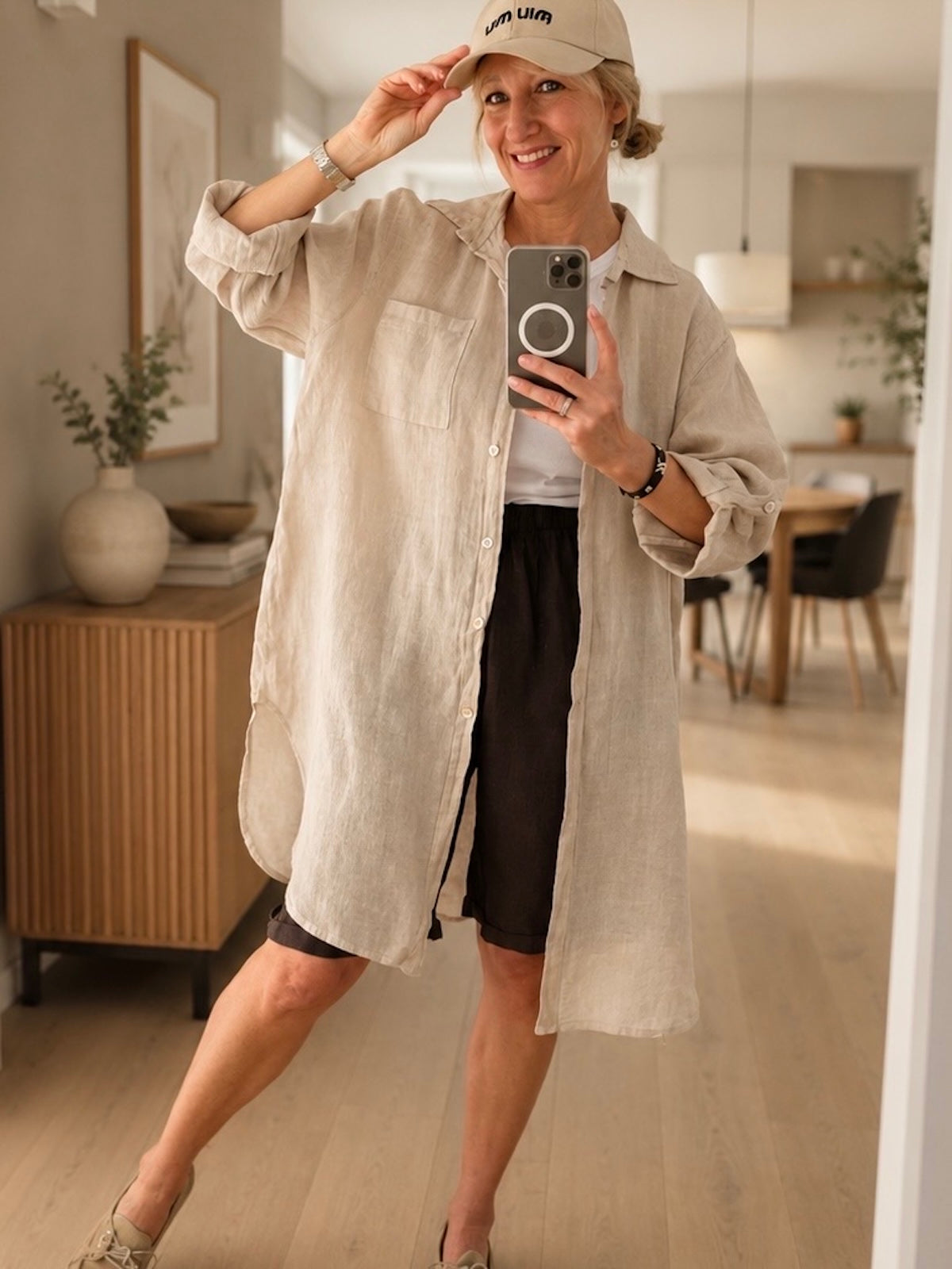 Kittel- oder Blusenkleid Bonny im hellen Farbton Stone aus strukturiertem Leinen-Gewebe, frontal präsentiert mit hochgekrempelten Ärmeln und Basecap im minimalistisch designten Store