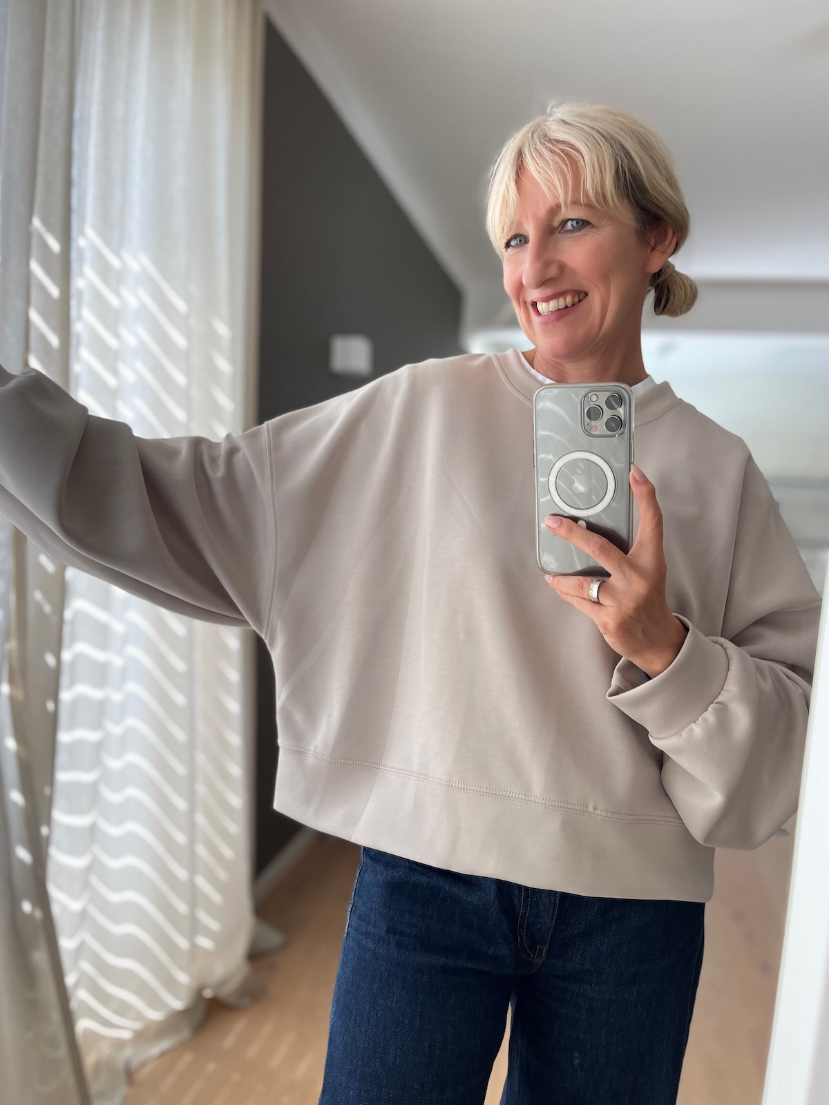 Blonde Frau zeigt den edlen, formstabilen Fall und die weiten Ärmel des beigen Boxy Sweatshirts Romy vor einem hellen Hintergrund.