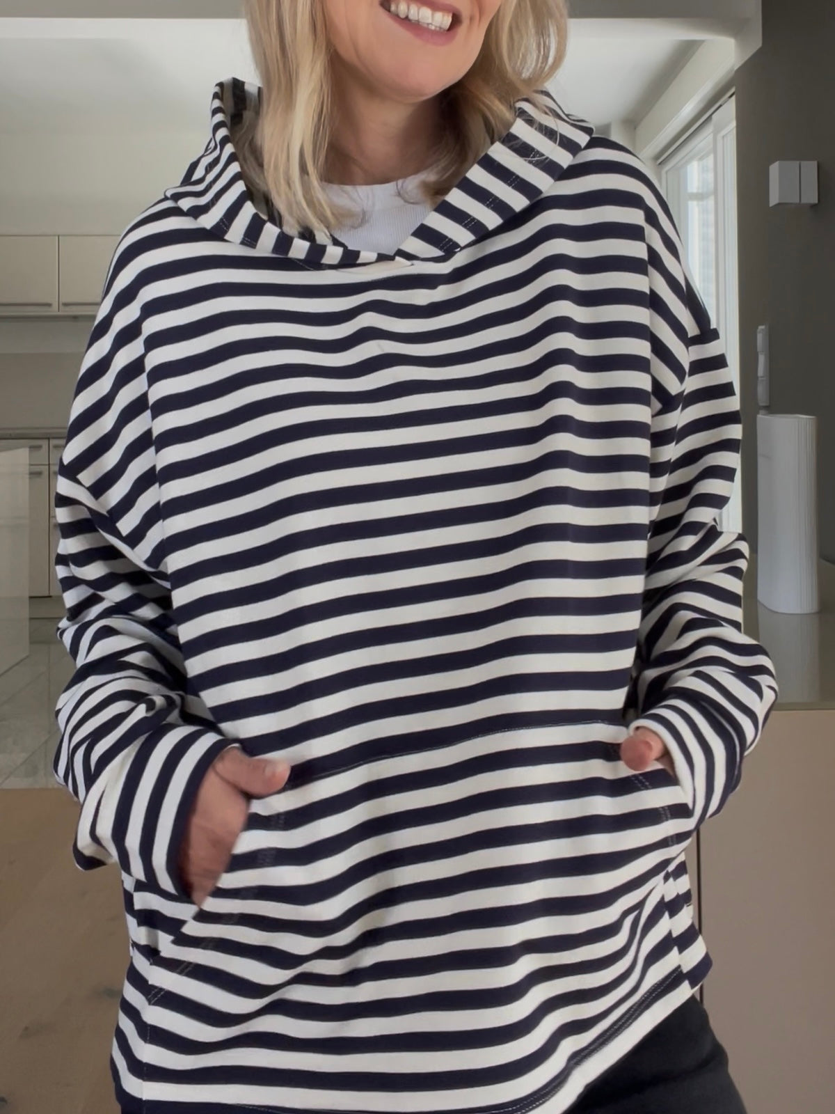Eine modebewusste Frau trägt den Hoodie Thea im Blau-Creme-Streifendesign, gestylt mit markanter Kapuze und aufgesetzter Fronttasche, der durch seinen strukturierten Fall besticht.