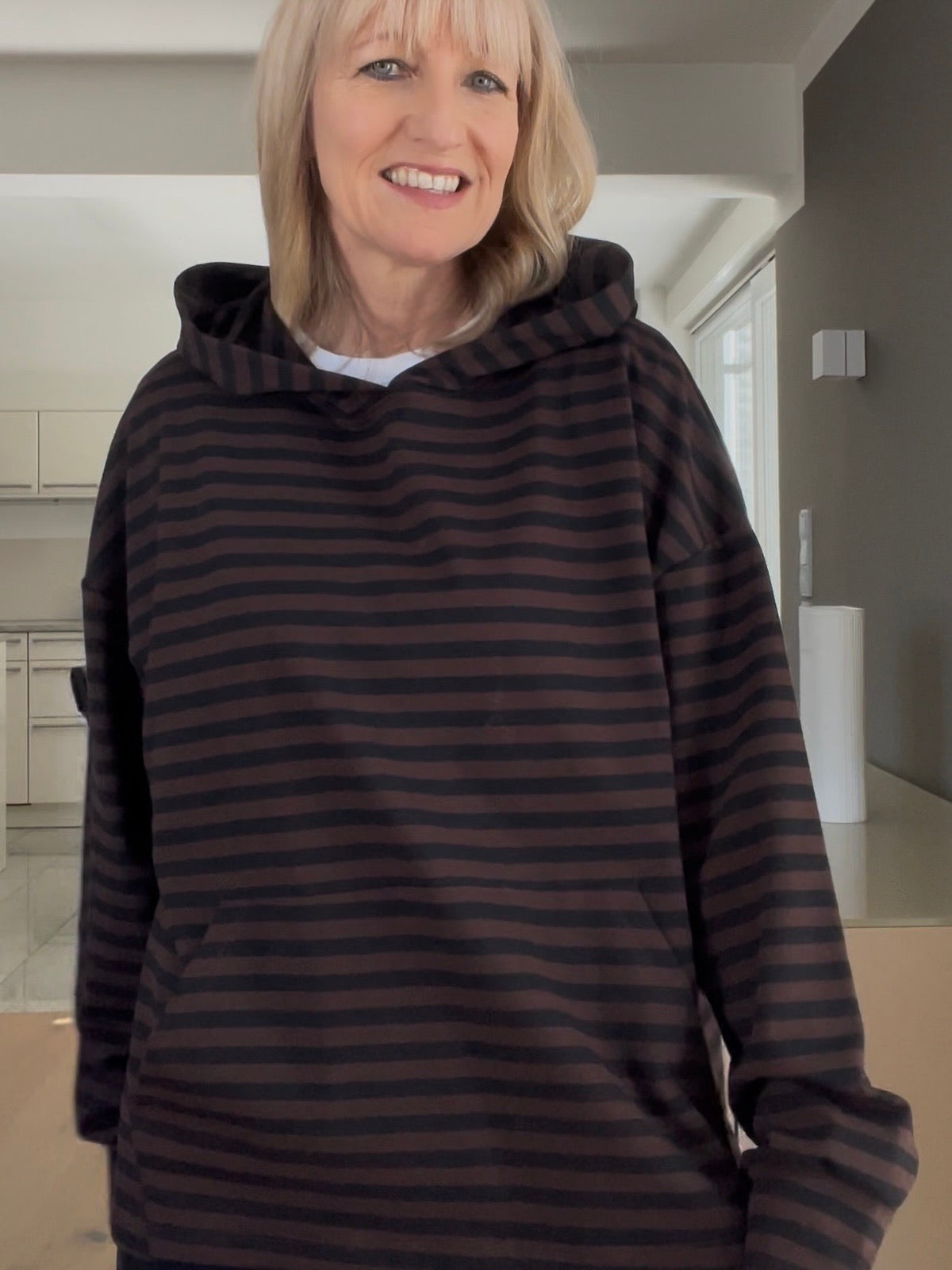 Eine modebewusste Frau trägt den Hoodie Thea im Schoko-Schwarz-Streifendesign, gestylt mit markanter Kapuze und aufgesetzter Fronttasche, der durch seinen strukturierten Fall besticht.
