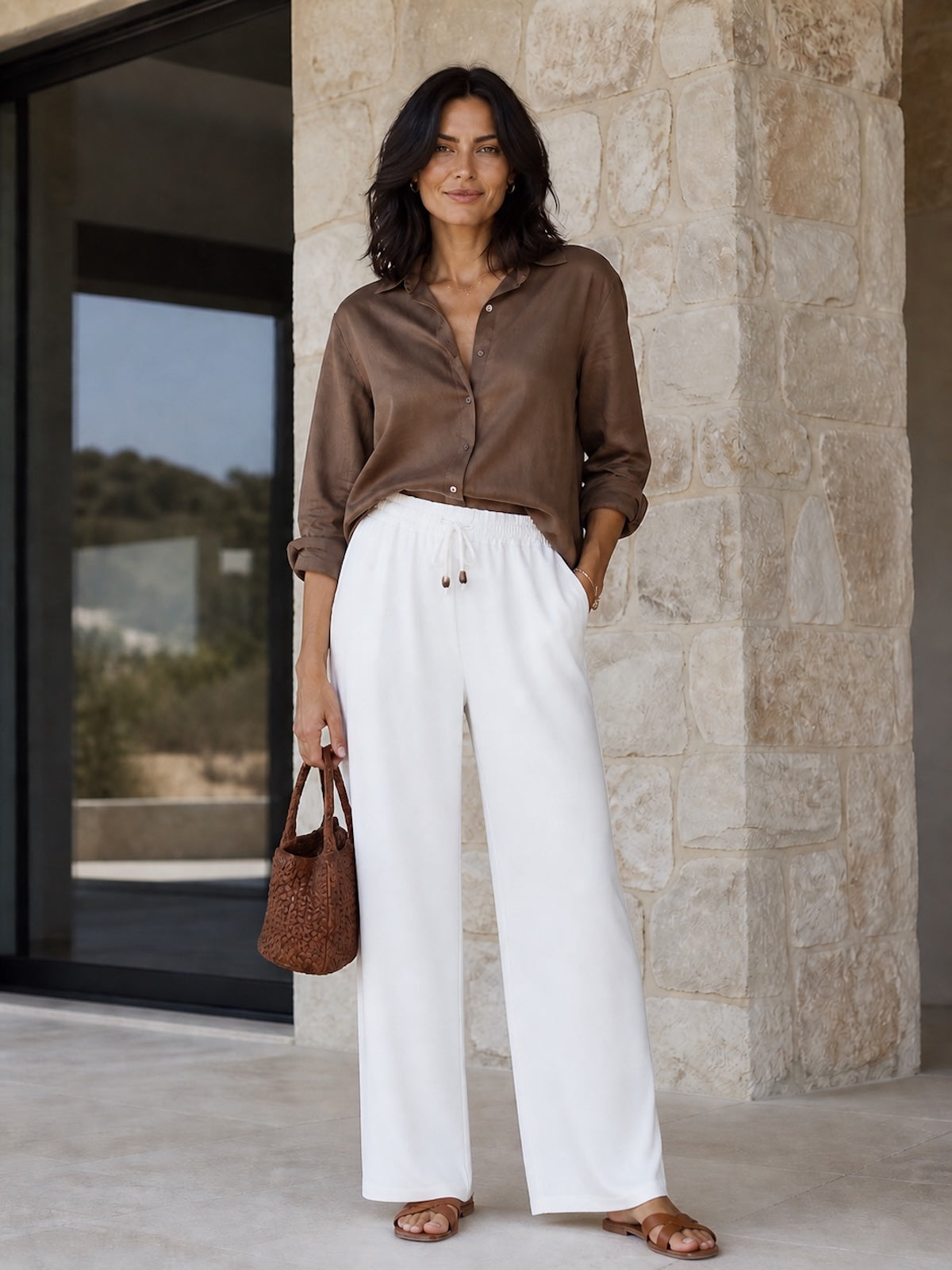 Brünettes Modell (1,70 m) trägt die weite, weiße Schlupfhose Tracy mit maritimen Tunnelzug und Eingrifftaschen, elegant kombiniert mit einer braunen Bluse, braunen Ledersandalen und einer geflochtenen Handtasche
