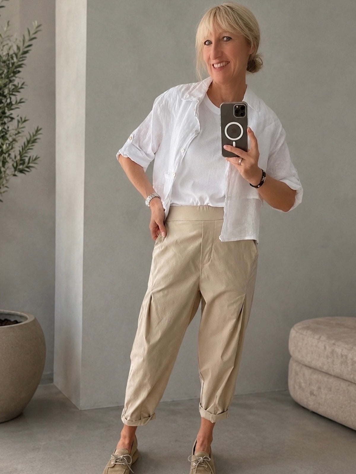 Frontansicht der sportlich-eleganten Hose Larissa in Stone an blondem Modell, kombiniert mit weißer Leinenbluse und beigen Wildleder-Loafers im minimalistischen Studio-Ambiente.