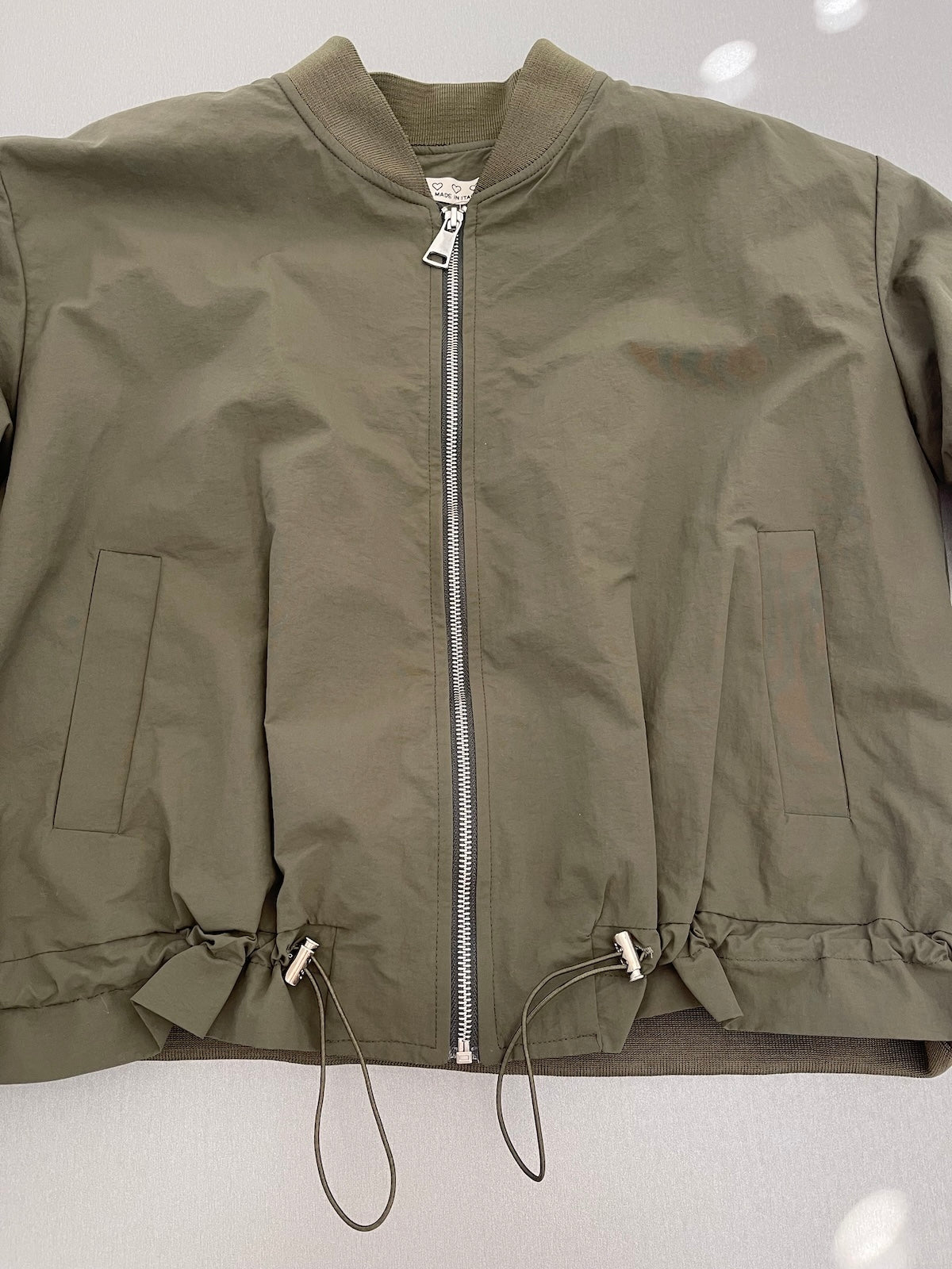 Grüne Damen-Bomberjacke Barbara Made in Italy mit silbernem Reißverschluss und verstellbarem Saum, flachliegend fotografiert.