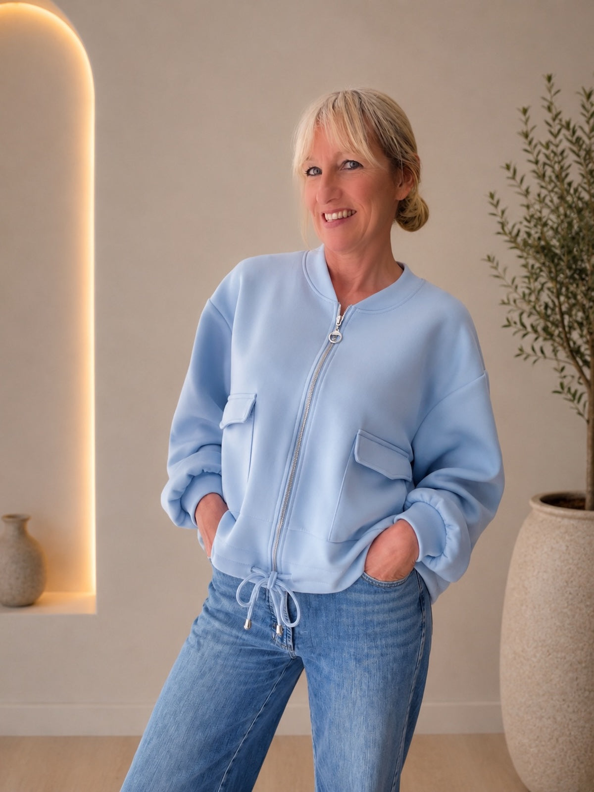 Lächelndes Model in einer sportlich-eleganten, hellblauen Blouson-Jacke Franzi mit Reißverschluss und Klappentaschen, lässig kombiniert mit einer blauen Jeans.