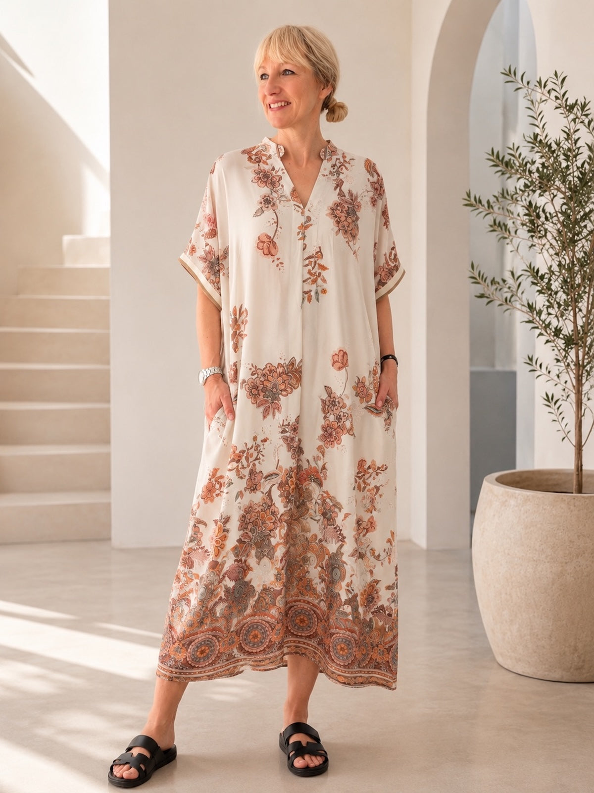 Frontansicht des eleganten Viskose-Seidenkleids Miranda in Creme-Floral an einem blonden Modell, kombiniert mit flachen schwarzen Sandalen vor einer hellen, modernen Loft-Kulisse.