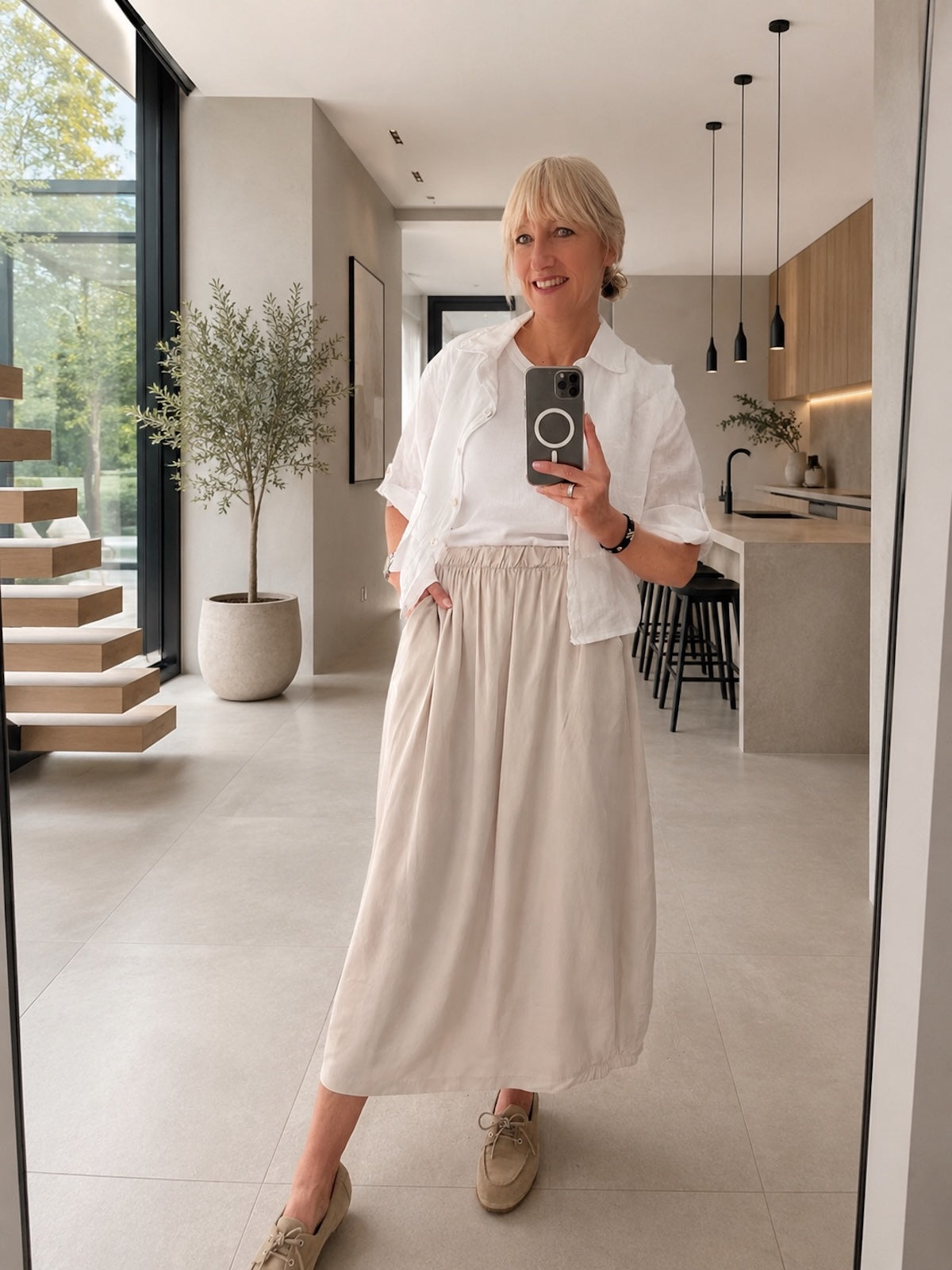 Leichtes weißes Damen Leinenhemd, offen getragen über einem cleanen T-Shirt zu einem fließenden, beigen Maxi-Rock für eine perfekt definierte Silhouette, Ganzkörperansicht im hellen Wohnbereich