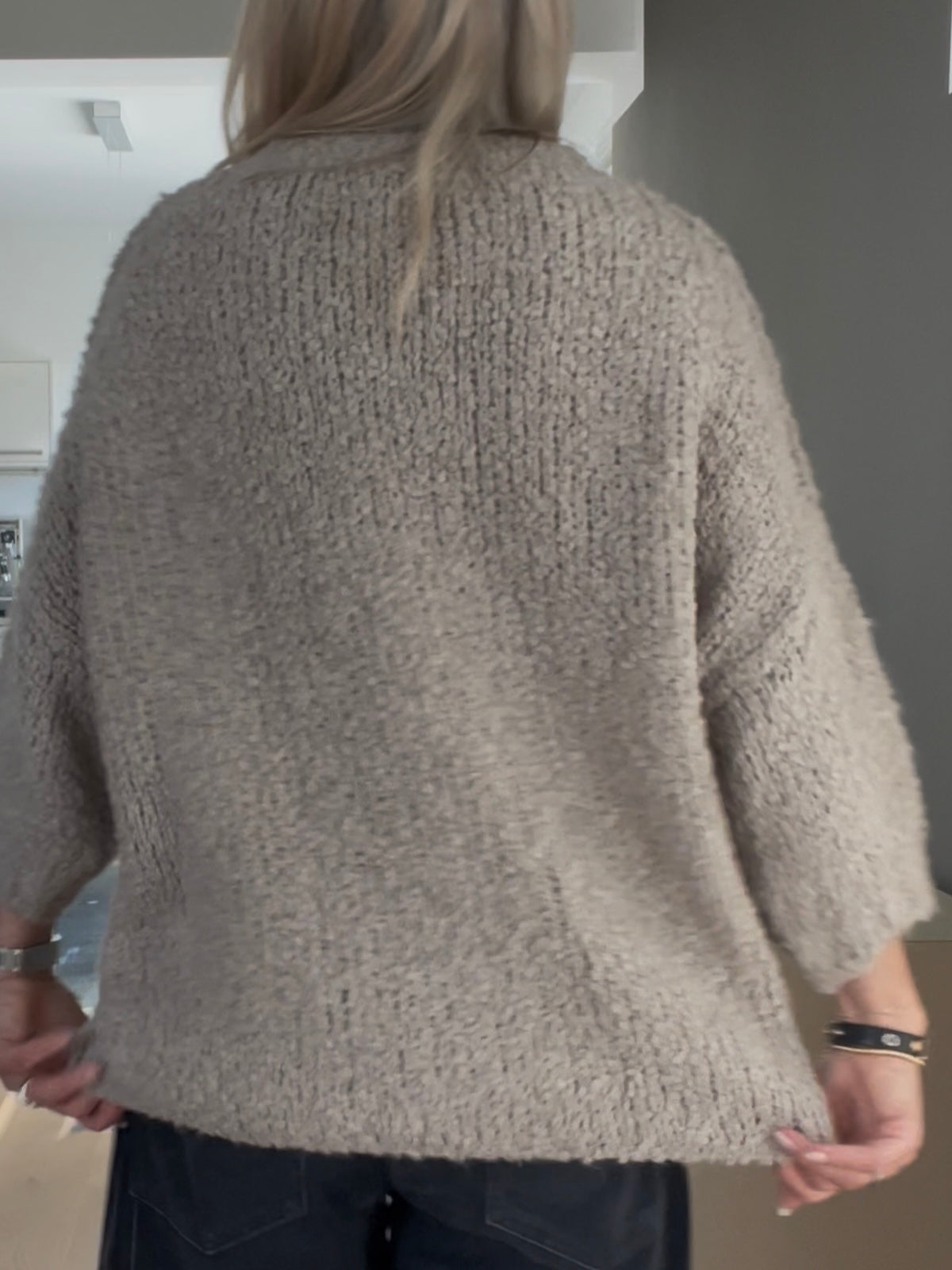 Rückansicht der Frau im greigefarbenen Pullover Jola, die das kompakte Maschenbild, den strukturierten Fall und die überschnittenen Schultern zeigt.