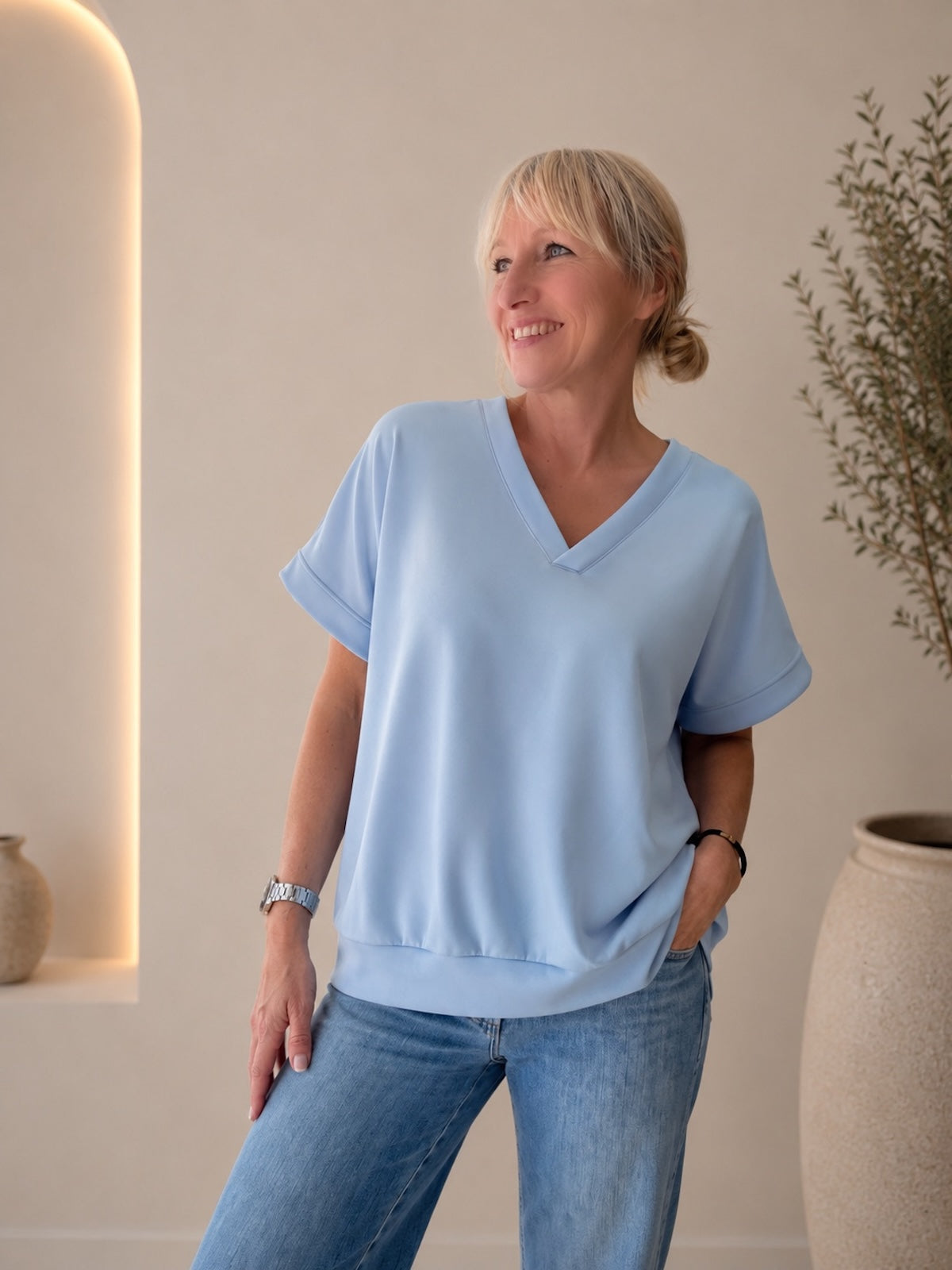 Lächelndes Model in dem eleganten hellblauen Kurzarm-Shirt Christine mit V-Ausschnitt und breitem Saumbündchen, kombiniert mit einer klassischen blauen Jeans.