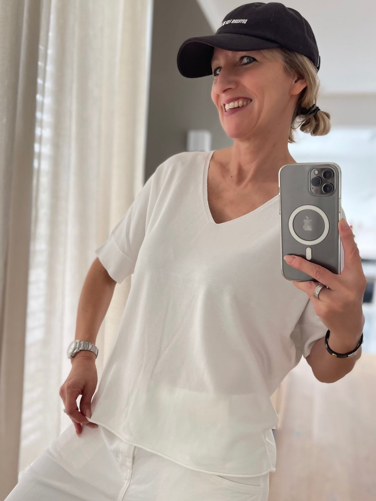 Silke von SILKE MODEN trägt das cremeweiße V-Ausschnitt Shirt Lisa im All-White-Look mit weißer Jeans und einer schwarzen Cap, entspanntes Spiegel-Selfie.