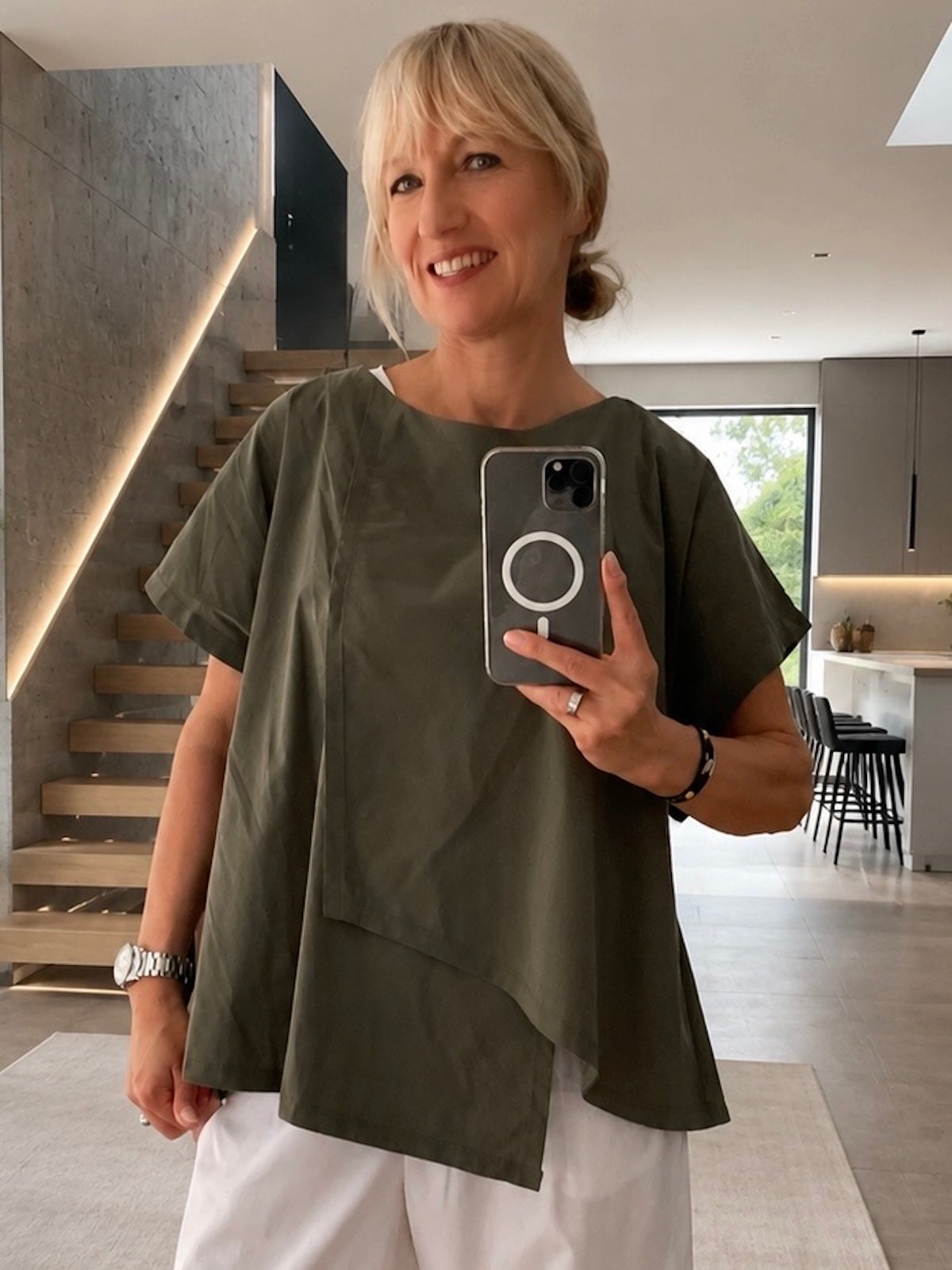 Exklusives Damen Shirt Marta in Khaki mit asymmetrischem Panel-Design, getragen von blondem Modell zu einer weißen Hose für eine sportlich-elegante Silhouette, Frontalansicht im modern gestalteten Interieur
