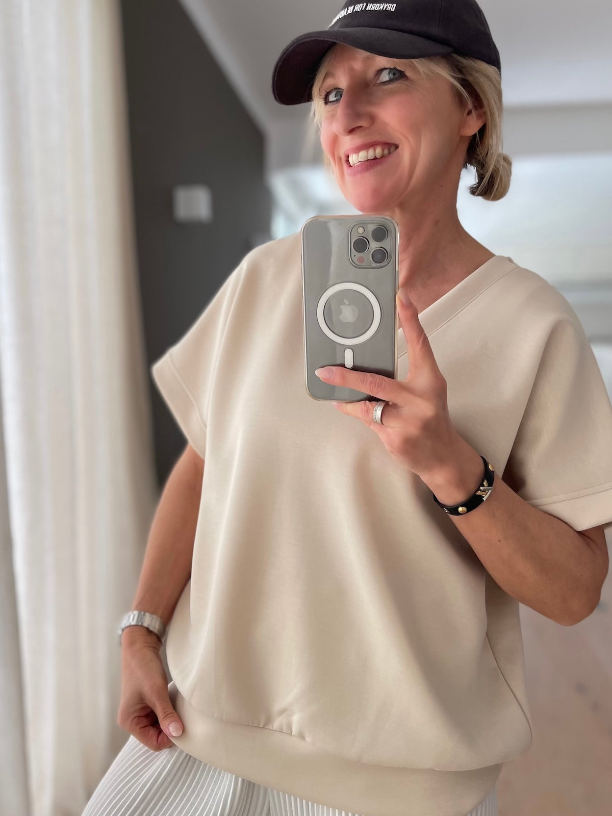 Blondes Modell (1,68 m) trägt das beigefarbene Kurzarm-Shirt Christine mit V-Ausschnitt und vorteilhaftem Saumbündchen, lässig gestylt zu einem weißen Plisseerock und dunkler Cap.