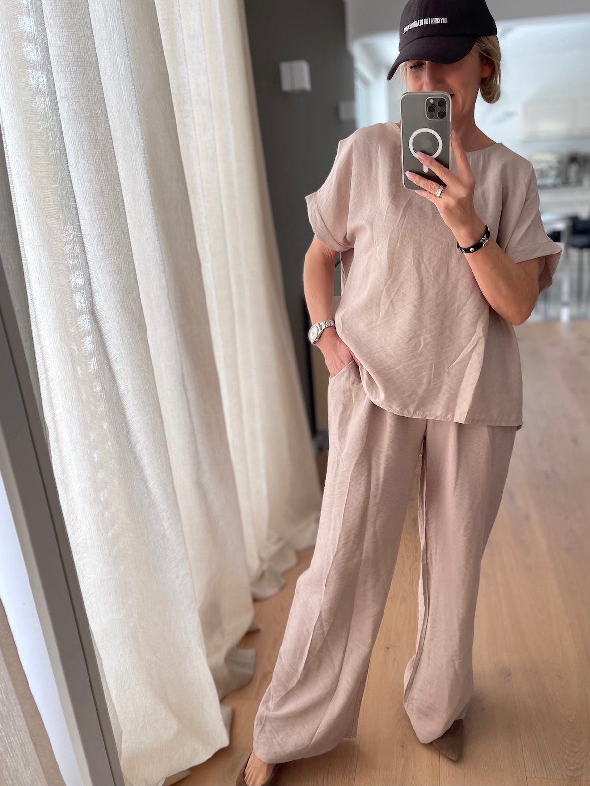 Blondes Modell trägt das fließende, taupefarbene Shirt Diana im lässigen Oversized-Fit, elegant kombiniert als Zweiteiler mit der farblich passenden Hose und einer schwarzen Cap.