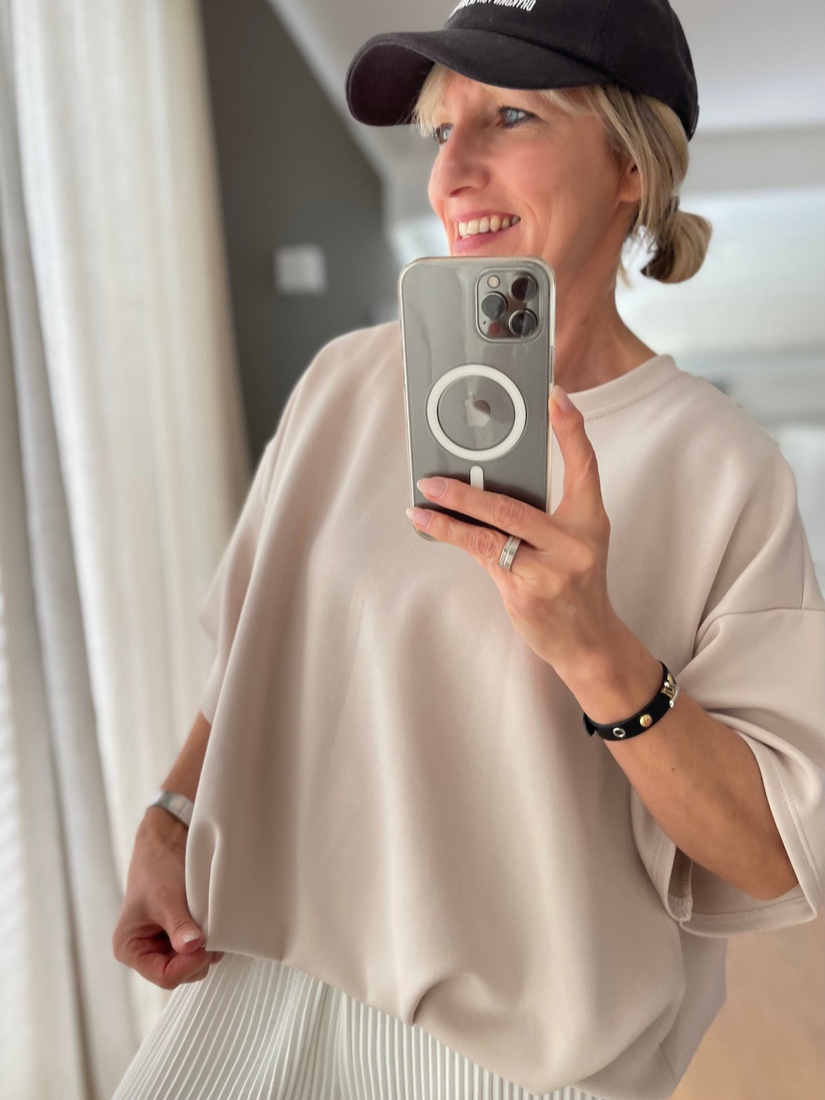 Blondes Modell präsentiert das glatte, hellbeige Oversized-Shirt Lilly mit Rundhalsausschnitt und weiten Ärmeln, sportlich kombiniert mit einem weißen Plisseerock und einer dunklen Cap.