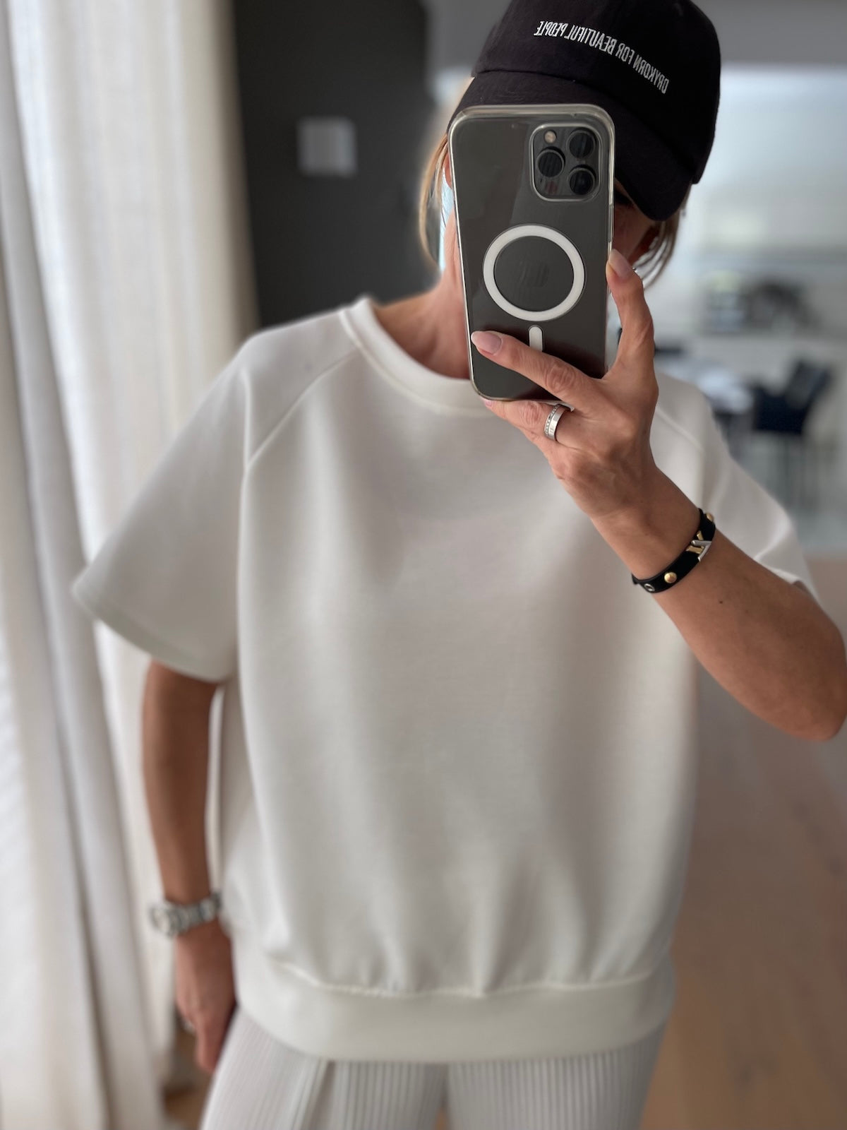 Blondes Modell trägt das strahlend weiße Shirt Nicole mit sportlichen Raglanärmeln und breitem Saumbündchen, lässig gestylt zu einem weißen Plissee-Unterteil und verdeckt durch das Smartphone beim Selfie.