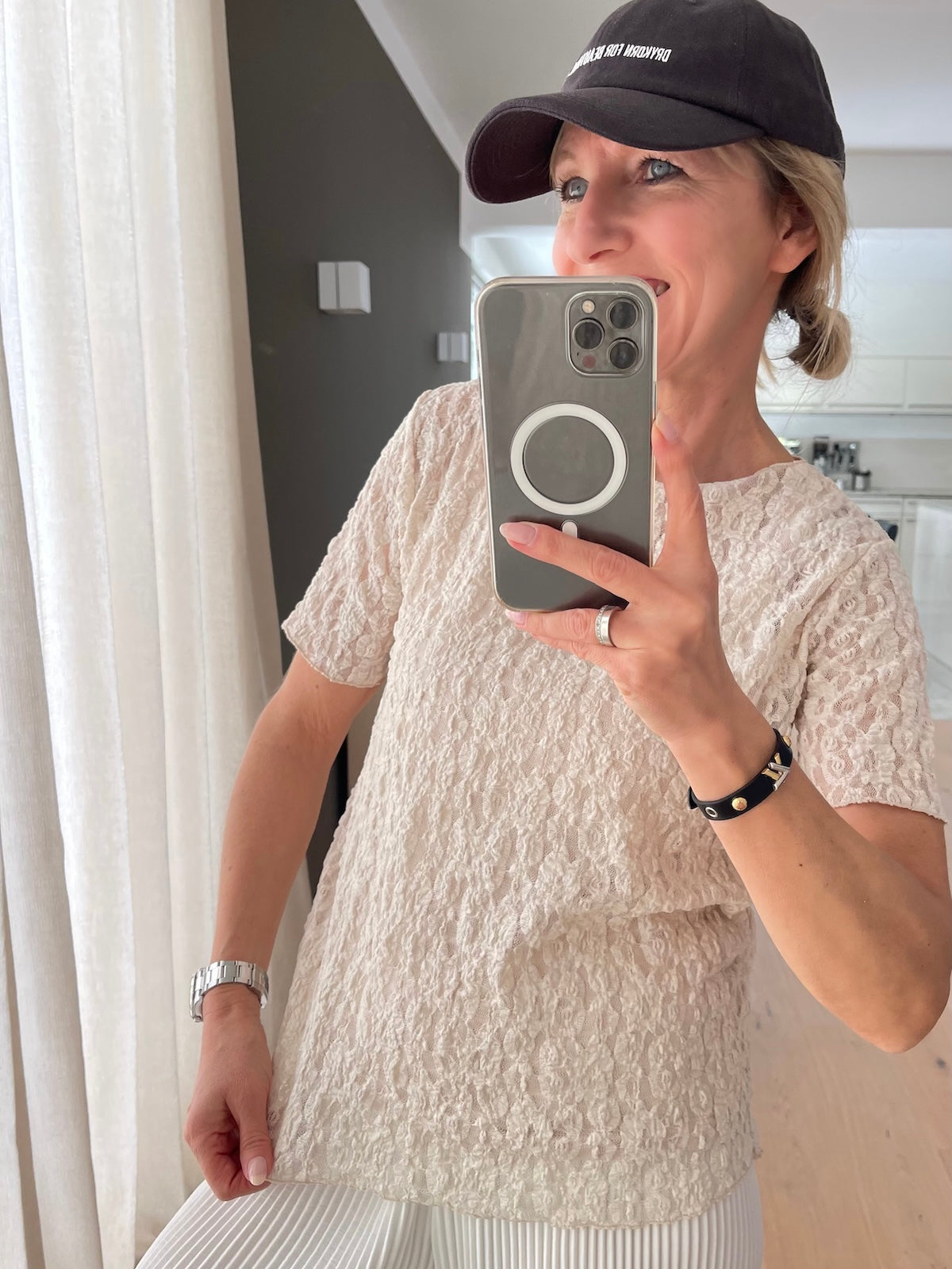Blondes Modell präsentiert das leicht transparente, beigefarbene Spitzenshirt Jade, unter dem ein farblich passendes Top getragen wird, elegant kombiniert mit einer weißen Plisseehose.