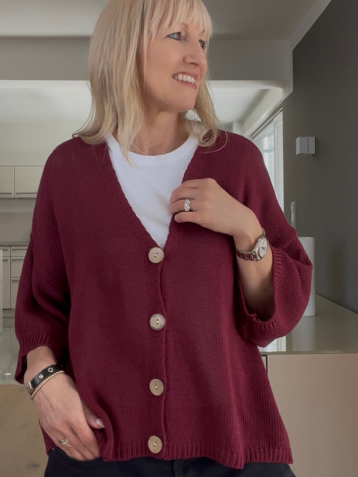 Eine modische Frau trägt den Strickcardigan Daisy in sattem Burgundi, gestylt mit tiefem V-Ausschnitt, markanten hellen Knöpfen und weiten Ärmeln im Boxy-Style, kombiniert mit einem weißen Shirt.