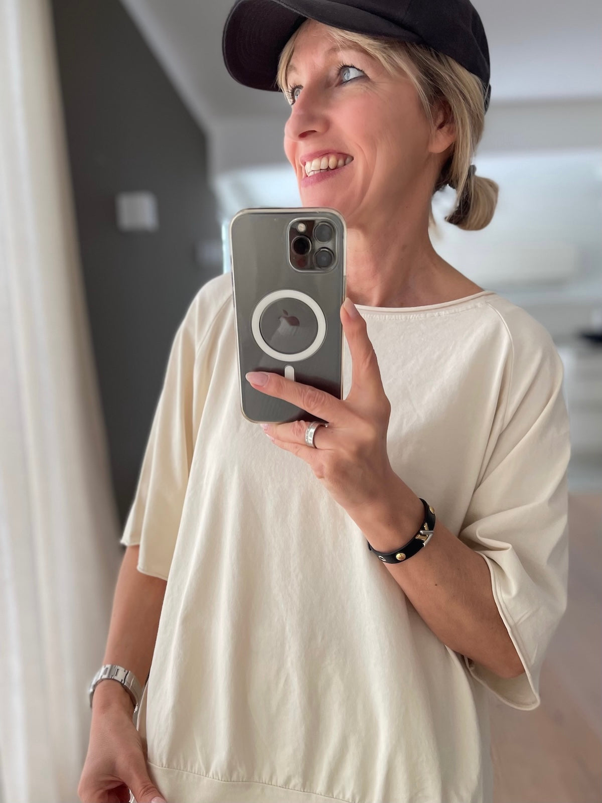 Lächelndes blondes Modell (1,68 m) trägt das cremefarbene Baumwoll-Shirt Mary mit weiten Raglanärmeln und figurfreundlichem Saumbündchen, sportlich gestylt zu einer hellen Plisseehose und schwarzer Cap.