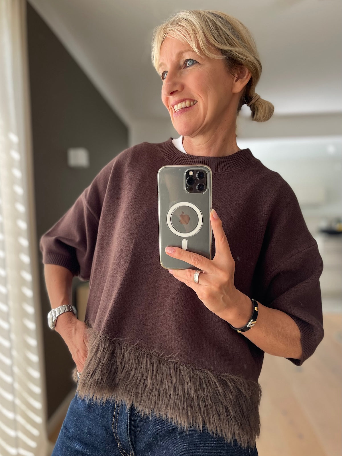 Blonde Frau posiert lächelnd und trägt den braunen Boxy Strickpullover Franzy mit Fransen am Saum, modisch kombiniert mit einer dunkelblauen High-Waist Jeans.