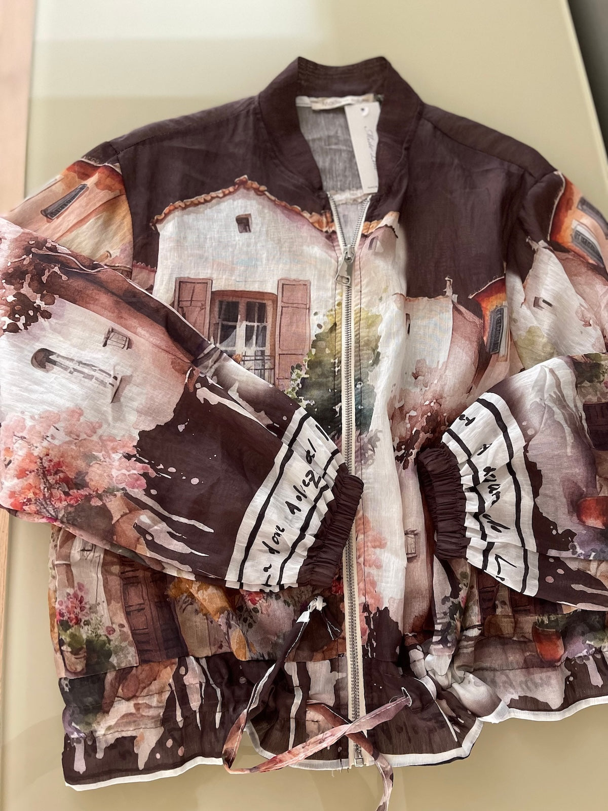 Flach liegende Detailaufnahme der ungefütterten Blouson-Jacke in Braun. Der malerische Art-Print, der silberfarbene Reißverschluss und das Zugband am Saum sind auf dem transparenten Stoff gut erkennbar.