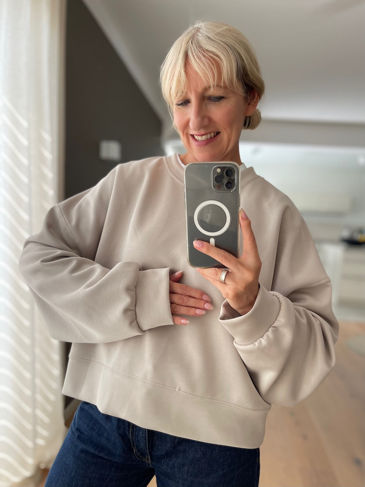 Blonde Frau präsentiert lächelnd das beige Boxy Sweatshirt Romy mit überschnittenen Schultern und trendigen Fledermausärmeln, modisch kombiniert mit einer dunkelblauen High-Waist Jeans.