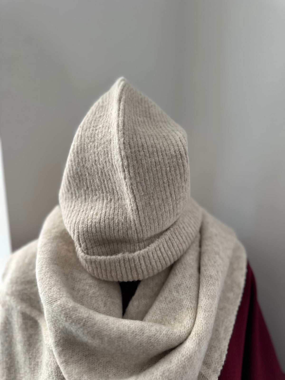 Beigefarbenes Accessoire-Set Alma, bestehend aus strukturiertem Dreieck-Schal und Mütze, stilvoll kombiniert auf einem bordeauxroten Oberteil.