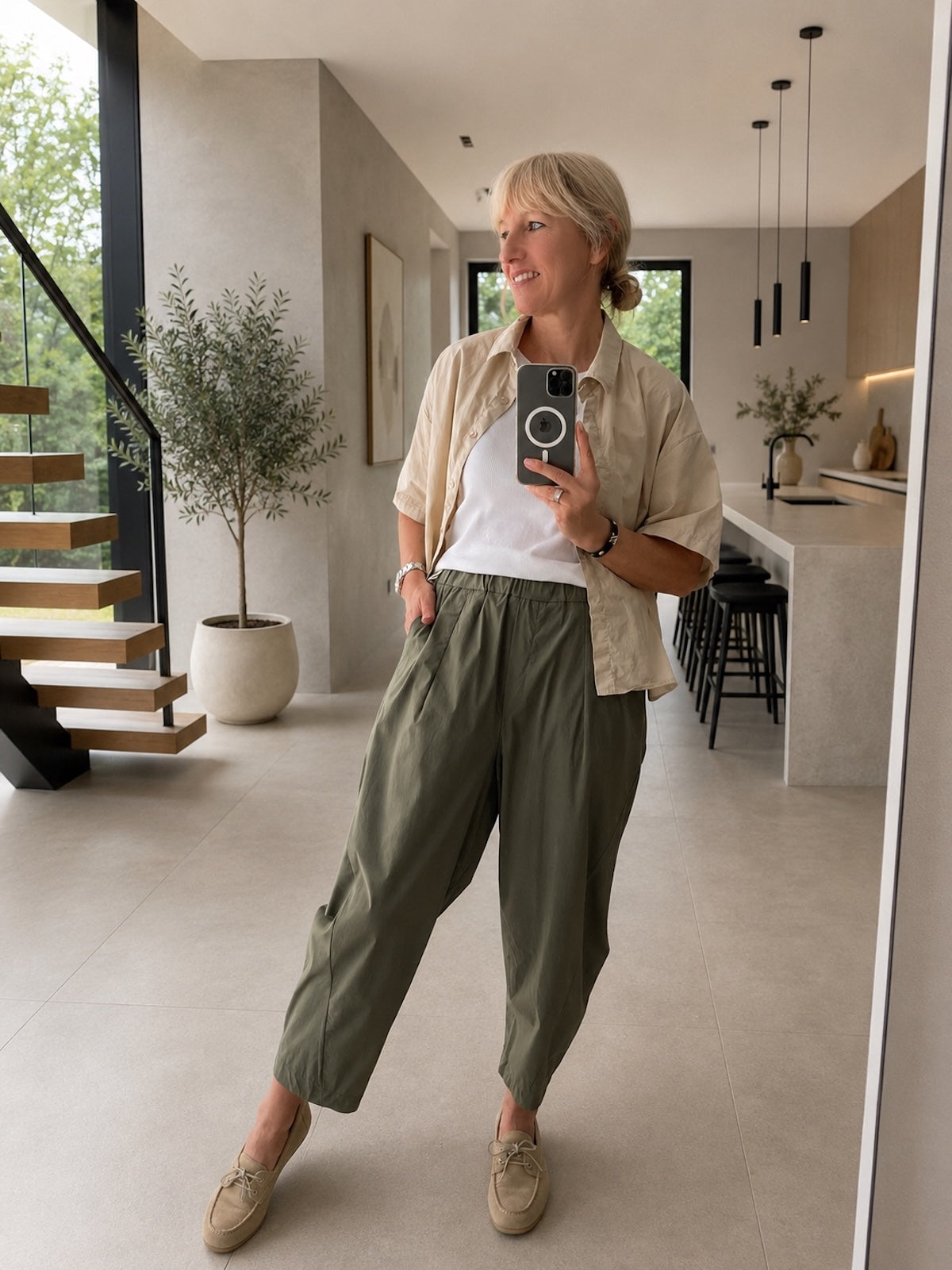 Ganzkörperansicht der sportlich-eleganten Hose Elisa in Khakigrün an einem blonden Modell, kombiniert mit einer offenen, beigen Kurzarmbluse und beigen Wildleder-Loafers im modernen Loft-Ambiente.