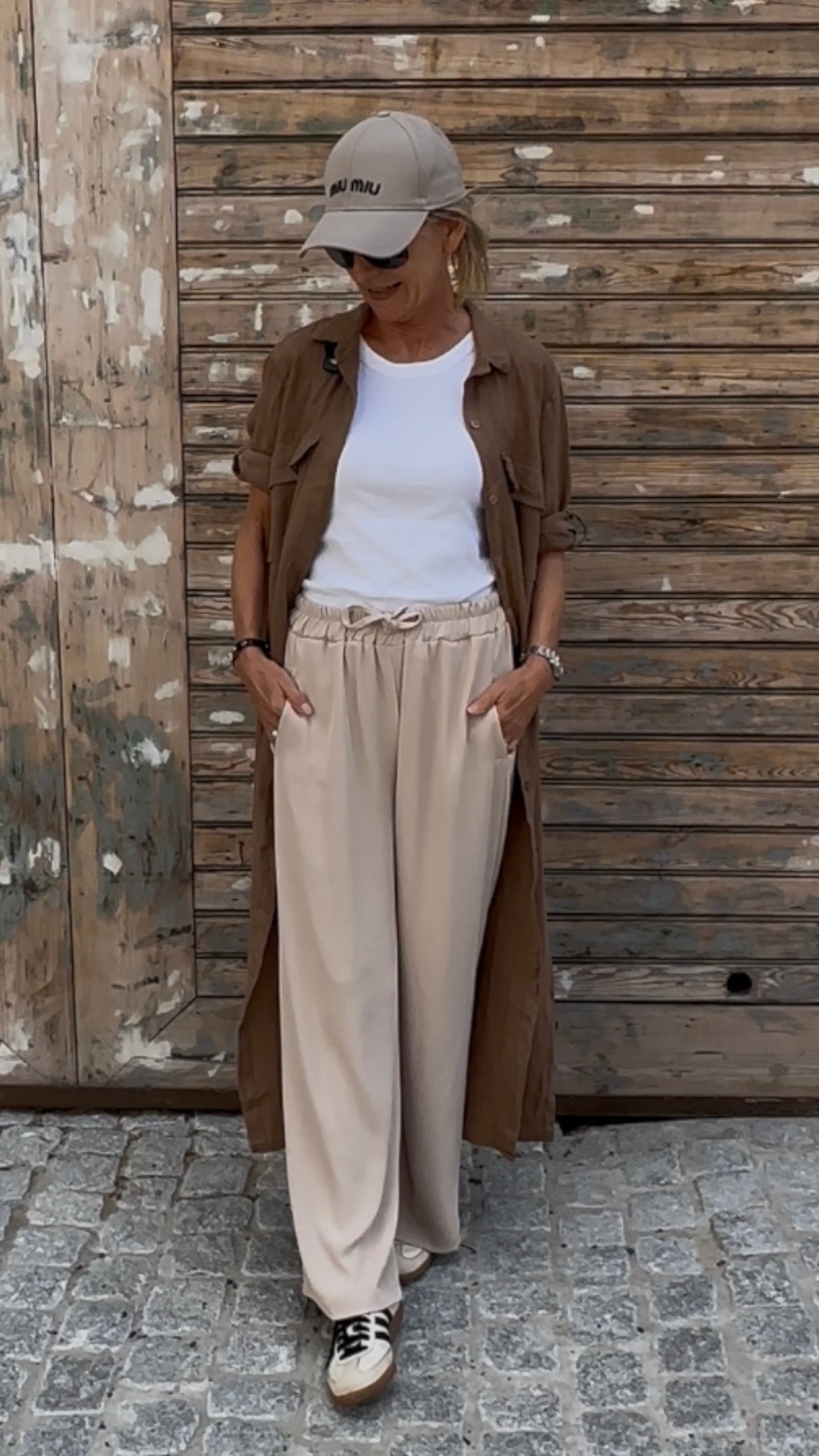 Model in der stone-farbenen Wide-Leg-Schlupfhose Tracy (Made in Italy), kombiniert mit Cap, Sonnenbrille und braunem Hemdkleid vor einem rustikalen Holztor.