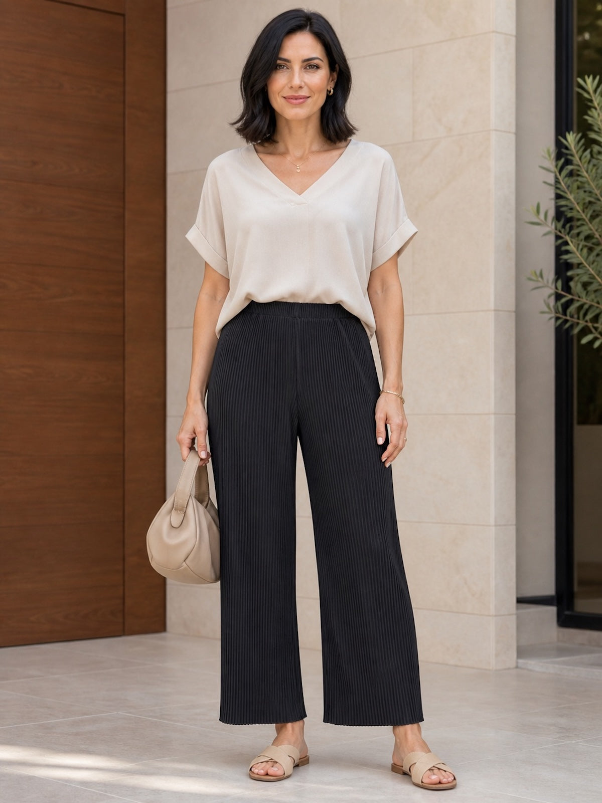 Brünettes Modell (1,70 m) trägt die knöchellange, schwarze Plissee-Hose Tony mit weitem Bein, elegant kombiniert mit einem beigefarbenen Kurzarm-Shirt, Ledersandalen und einer beigen Handtasche.
