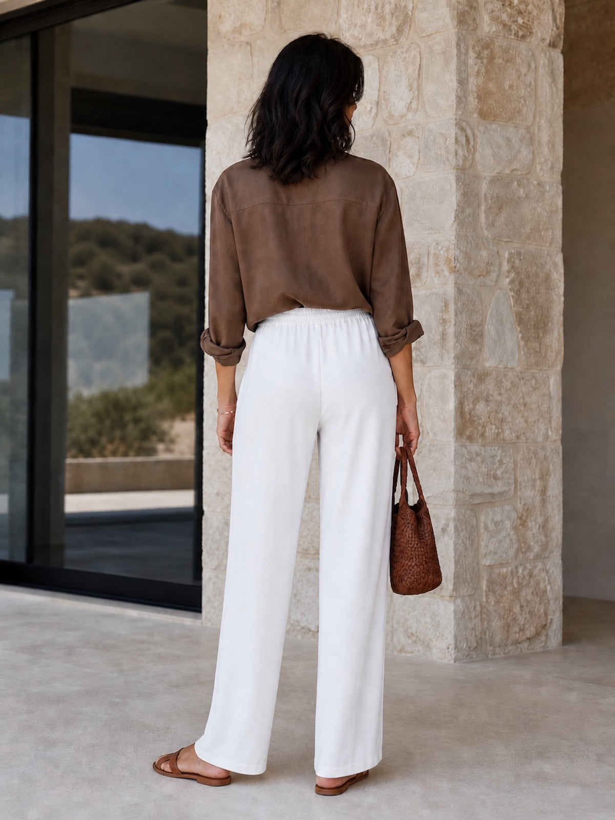 Rückansicht der weißen, fließenden Hose Tracy. Das Modell präsentiert den lockeren Fall des weiten Beins, lässig kombiniert mit einer braunen Bluse und braunen Accessoires auf einer Steinterrasse.