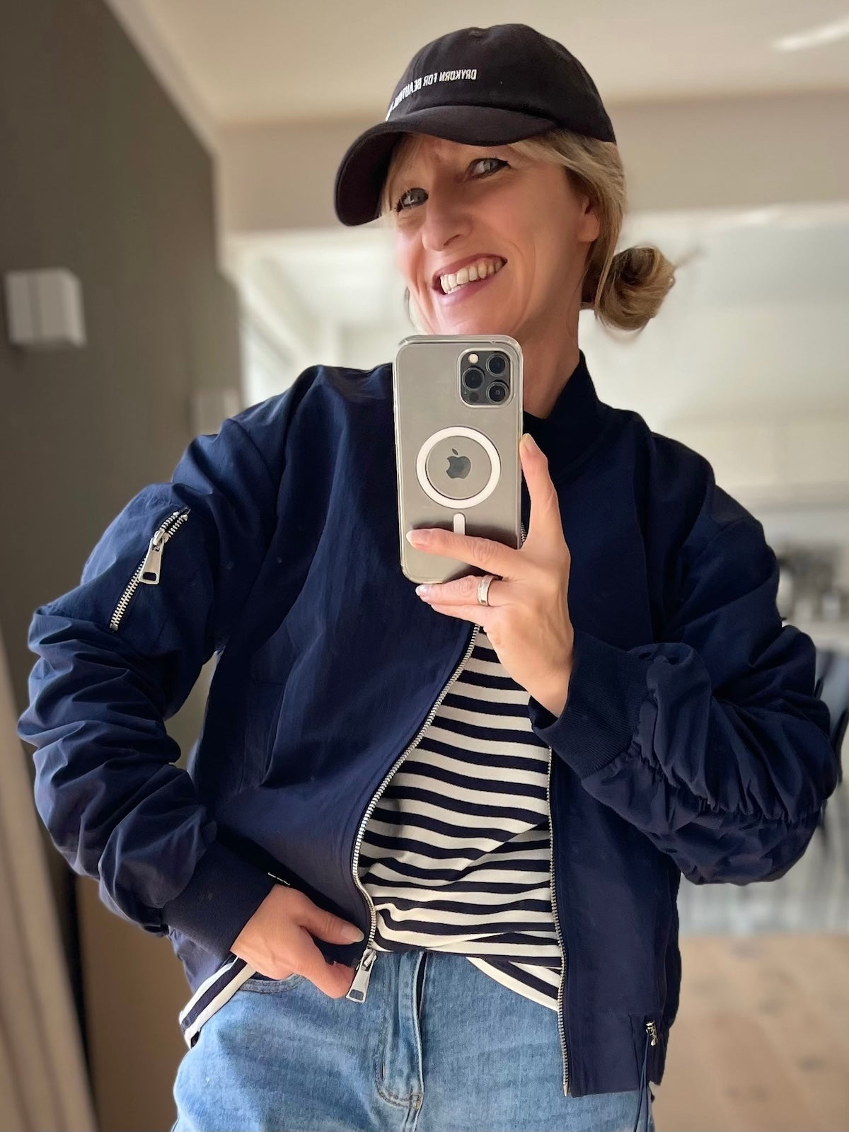 Lächelndes Model trägt die dunkelblaue Damen-Bomberjacke Barbara aus Nylon mit silbernem Reißverschluss und Tunnelzug am Saum, kombiniert mit einem gestreiften Shirt und einer Jeans.