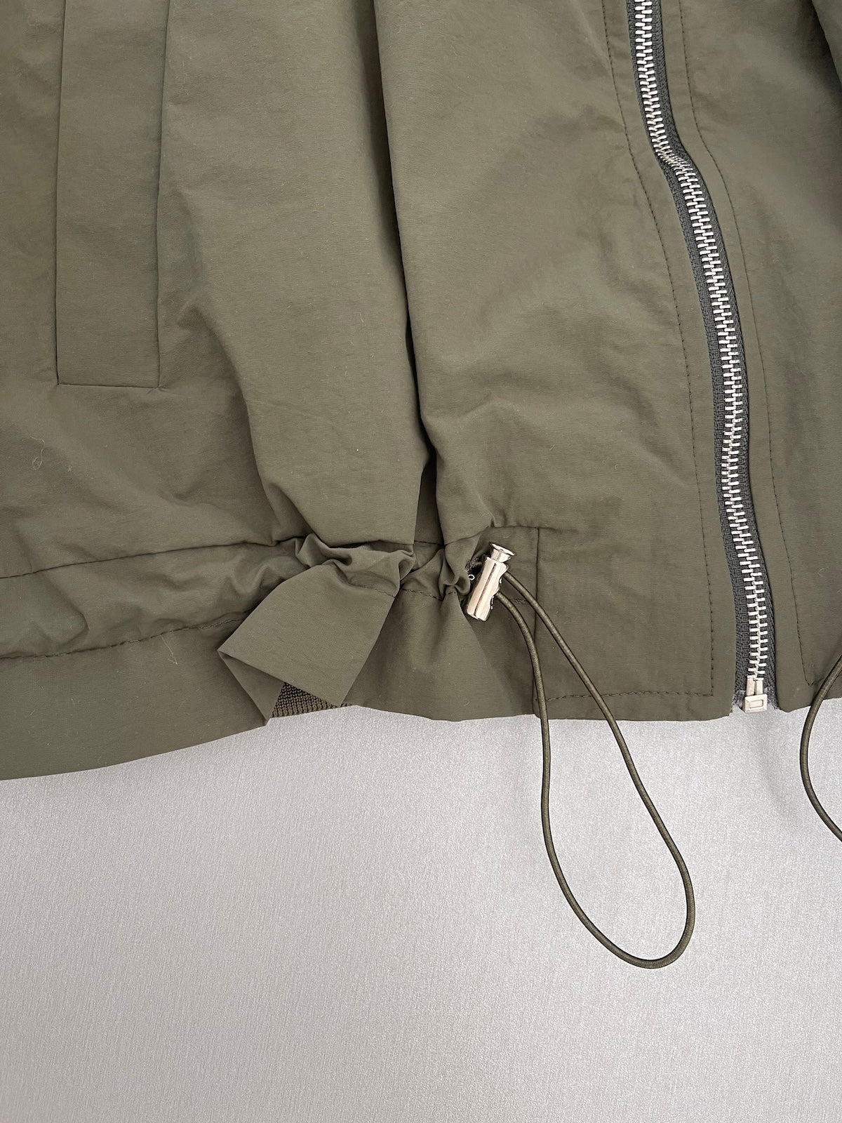 Detailaufnahme von Kordelzug und silbernem Stopper am gerafften Saum der grünen Übergangsjacke Barbara.