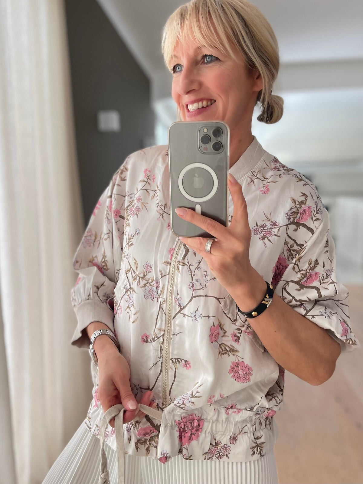 Blondes Modell trägt die cremeweiße Leichtjacke Rose mit zartrosa Blumenmuster, lässig geschnitten mit Reißverschluss und gerafftem Tunnelzug am Saum.