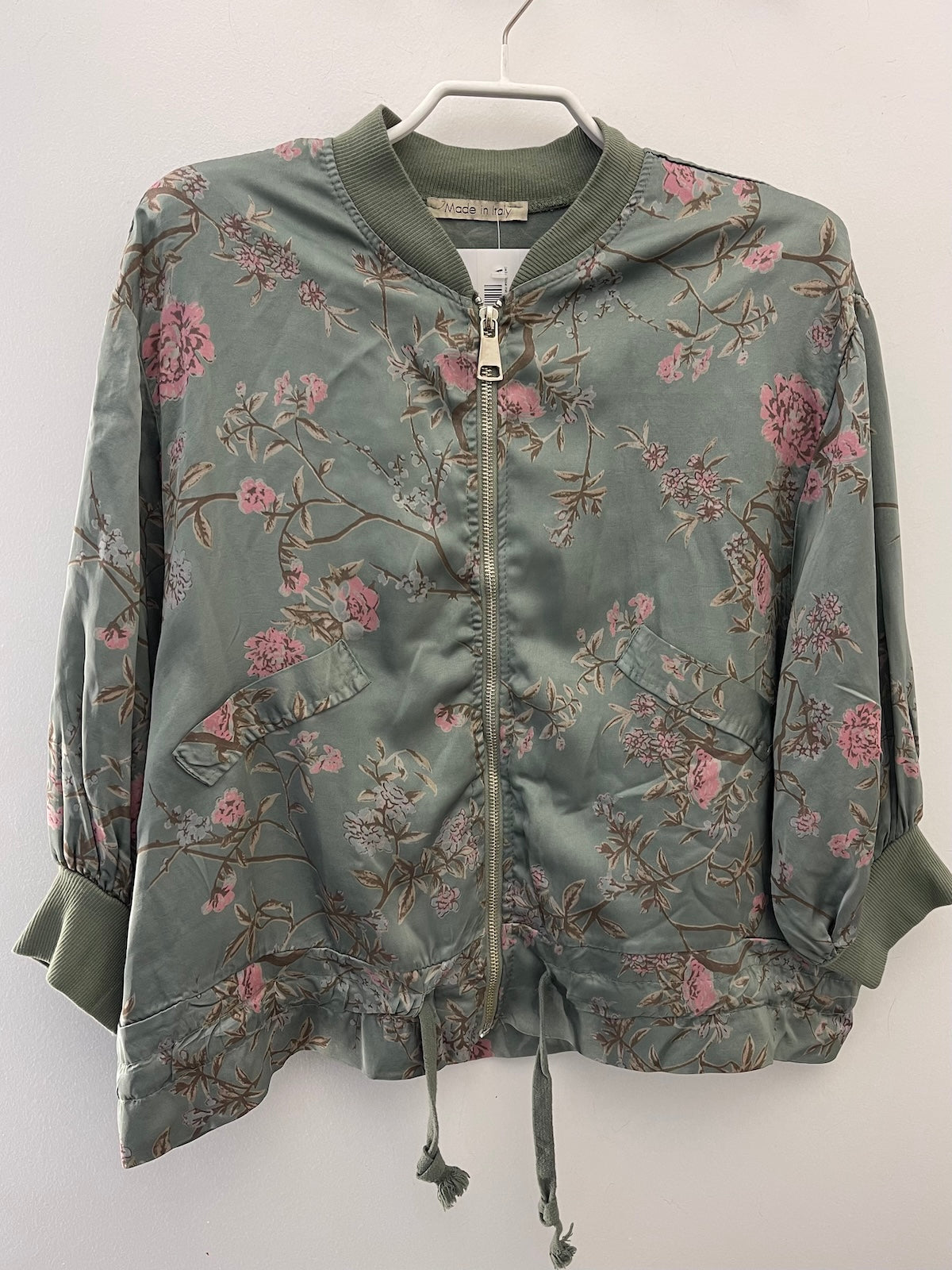 Auf einem weißen Kleiderbügel hängende Detailansicht der grünen Viskose-Jacke Rose mit floralem Print. Der silberfarbene Reißverschluss, der Rippstrick-Kragen und die Kordel am Saum sind gut sichtbar.