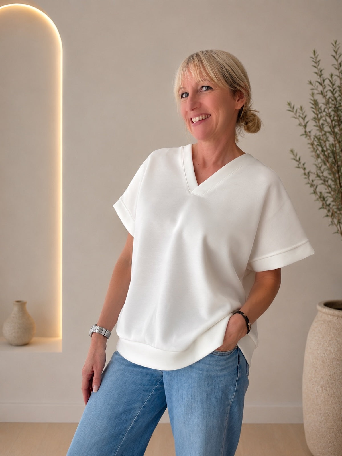 Lächelndes Model in dem eleganten weißen Kurzarm-Shirt Christine mit V-Ausschnitt und breitem Saumbündchen, kombiniert mit einer klassischen blauen Jeans.