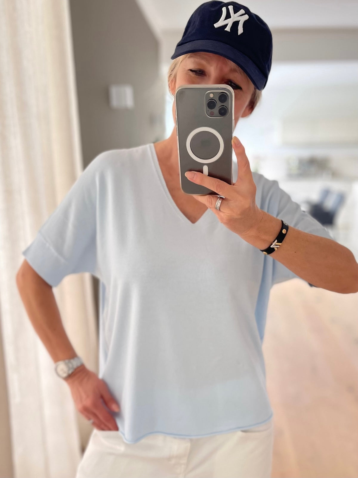 Silke von SILKE MODEN trägt das hellblaue V-Ausschnitt Shirt Lisa, lässig kombiniert mit einer weißen Jeans und einer dunkelblauen NY-Cap.