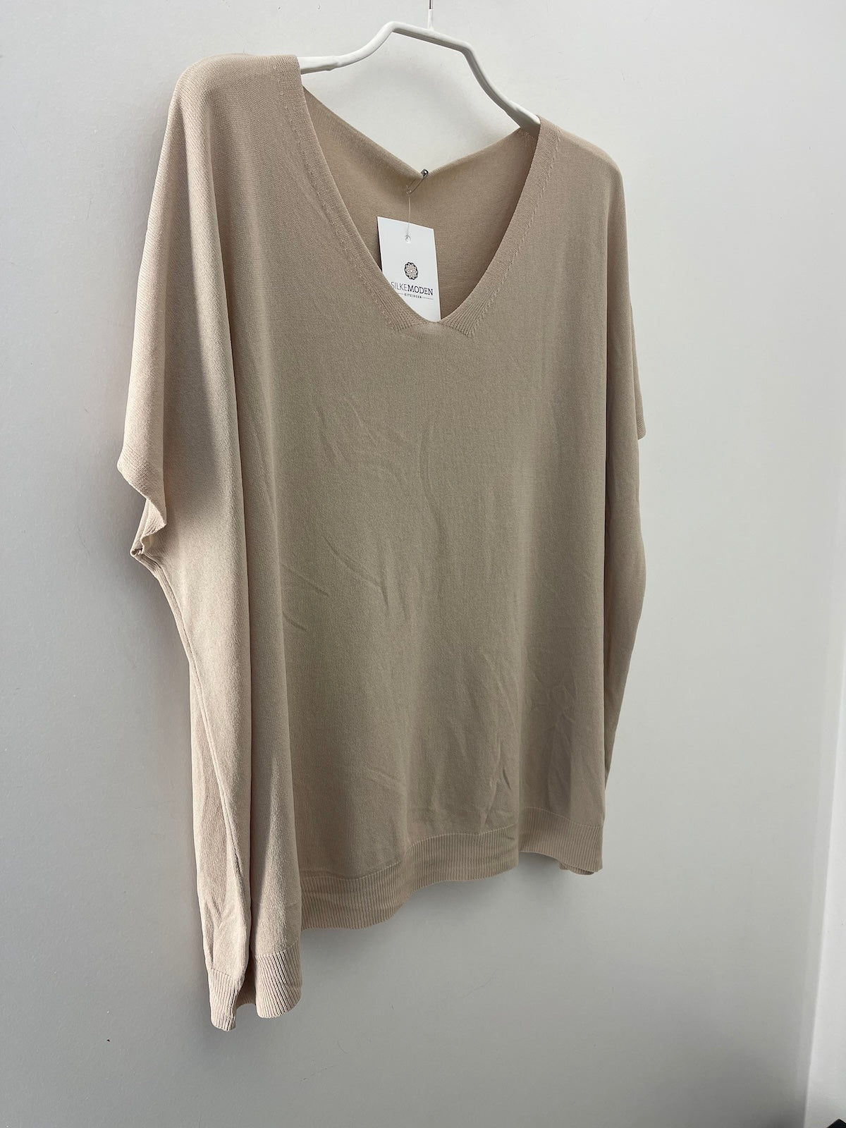 Eine am Bügel hängende Produktaufnahme des Damen-T-Shirts Modell Maria in der Farbe Beige. Das Shirt ist ordentlich auf einem neutralen Hintergrund präsentiert, um den lockeren Schnitt, den V-Ausschnitt und die Struktur des Stoffes detailliert darzustellen.