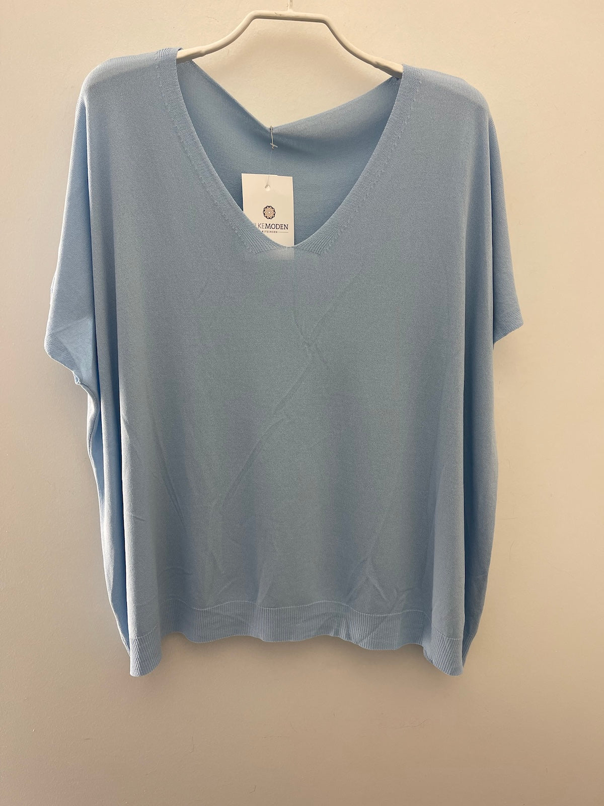 Eine am Bügel hängende Produktaufnahme des Damen-T-Shirts Modell Maria in der Farbe Hellblau. Das Shirt ist ordentlich auf einem neutralen Hintergrund präsentiert, um den lockeren Schnitt, den V-Ausschnitt und die Struktur des Stoffes detailliert darzustellen.
