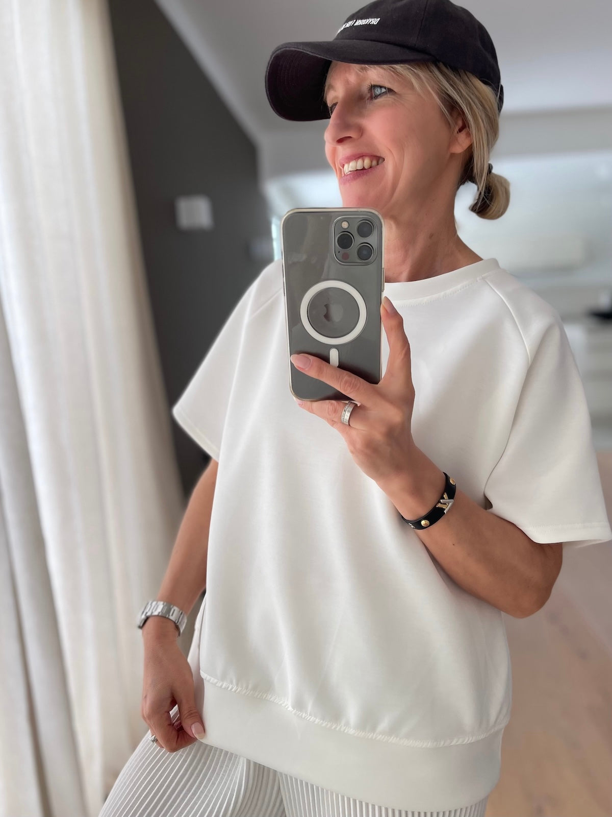 Lächelndes blondes Modell (1,68 m) im Spiegel-Selfie präsentiert den lockeren Fall und das cleane Design des weißen Kurzarm-Shirts Nicole, kombiniert mit einer schwarzen Cap.