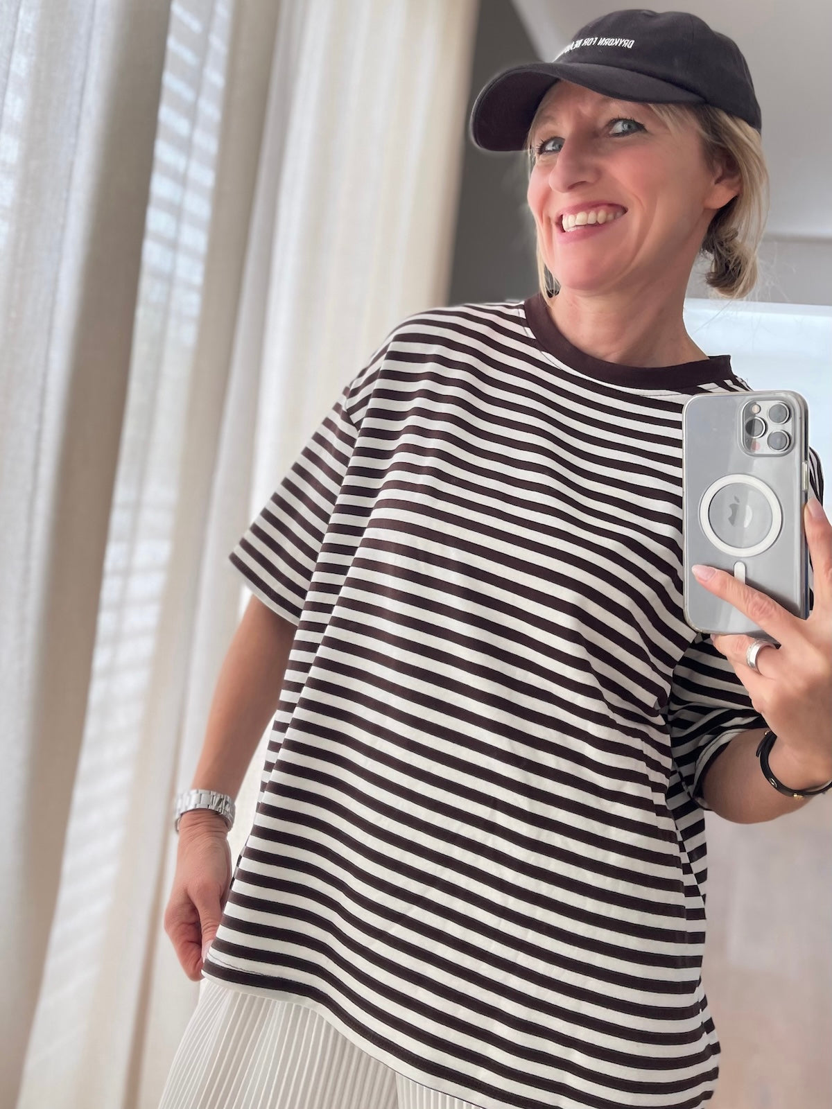 Blondes Modell (1,68 m) trägt das braun-weiß gestreifte Oversized-Shirt Nora aus weicher Viskose, sportlich kombiniert mit einem weißen Plisseerock und einer dunklen Cap.