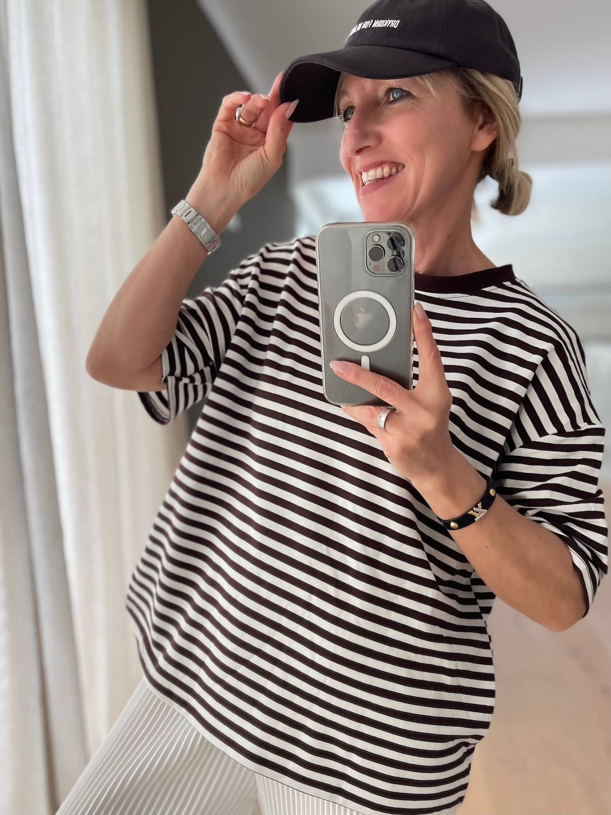 Lächelndes blondes Modell richtet ihre Kappe, trägt das fließende, gestreifte Kurzarm-Shirt Nora in Braun und Weiß, perfekt für einen sportlich-eleganten Sommerlook.