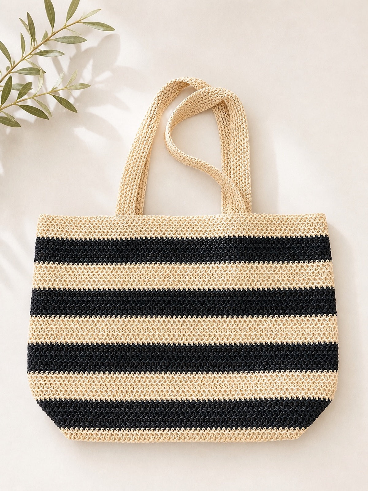 Ein flach liegender, geflochtener Sommer-Shopper namens Zoe im maritimen Streifen-Design in Beige und Schwarz. Die Tasche aus Bast hat zwei Tragehenkel und wird stilvoll auf einem hellen Hintergrund mit einem feinen Olivenzweig präsentiert.