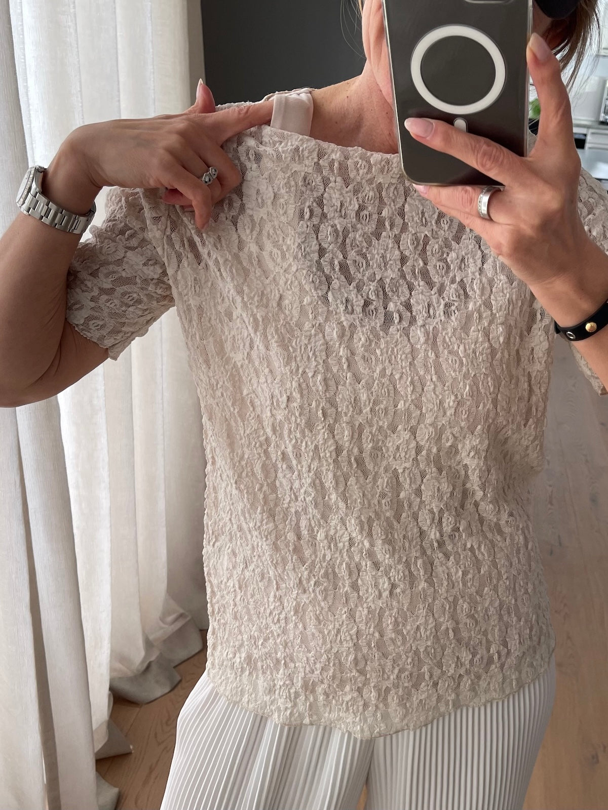 Lächelndes blondes Modell im Spiegel-Selfie mit dunkler Cap, das romantisch strukturierte, beigefarbene Spitzenshirt Jade fällt leicht und feminin.