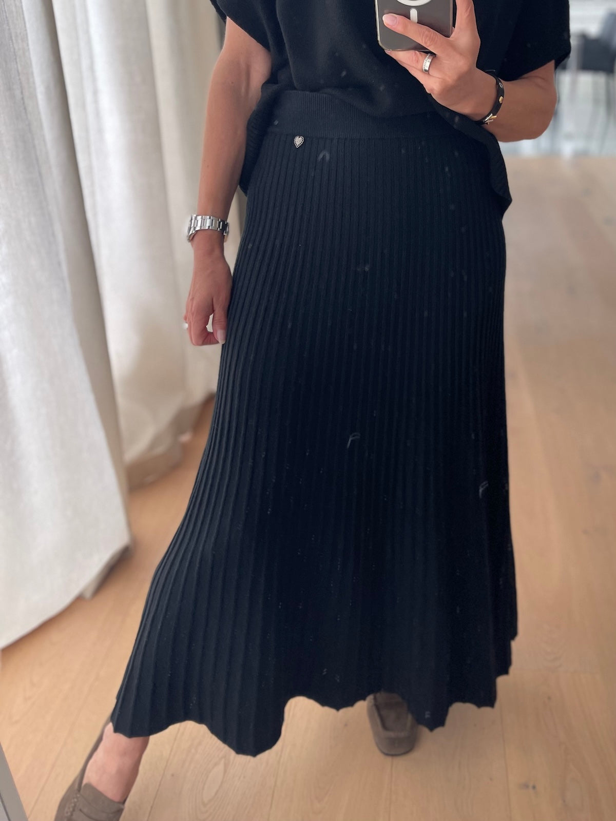 Modebewusste Frau trägt den schwarzen Strickrock Elise in feiner Plissee-Optik mit einer klar definierten Silhouette, perfekt abgestimmt auf den exakt passenden, schwarzen Strickpullunder Edda.