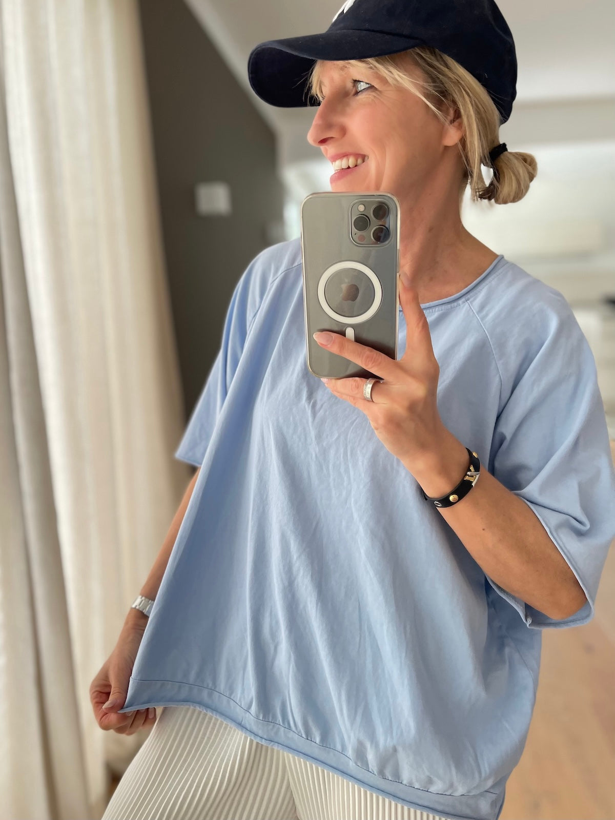 Lächelndes blondes Modell (1,68 m) trägt das hellblaue Baumwoll-Shirt Mary mit weiten Raglanärmeln und figurfreundlichem Saumbündchen, lässig gestylt zu einer weißen Plisseehose und einer dunkelblauen Cap.